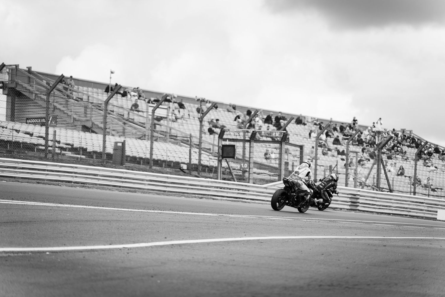 Black and white photo of a motorcycle racing on a circuit track with spectators in the grandstand.