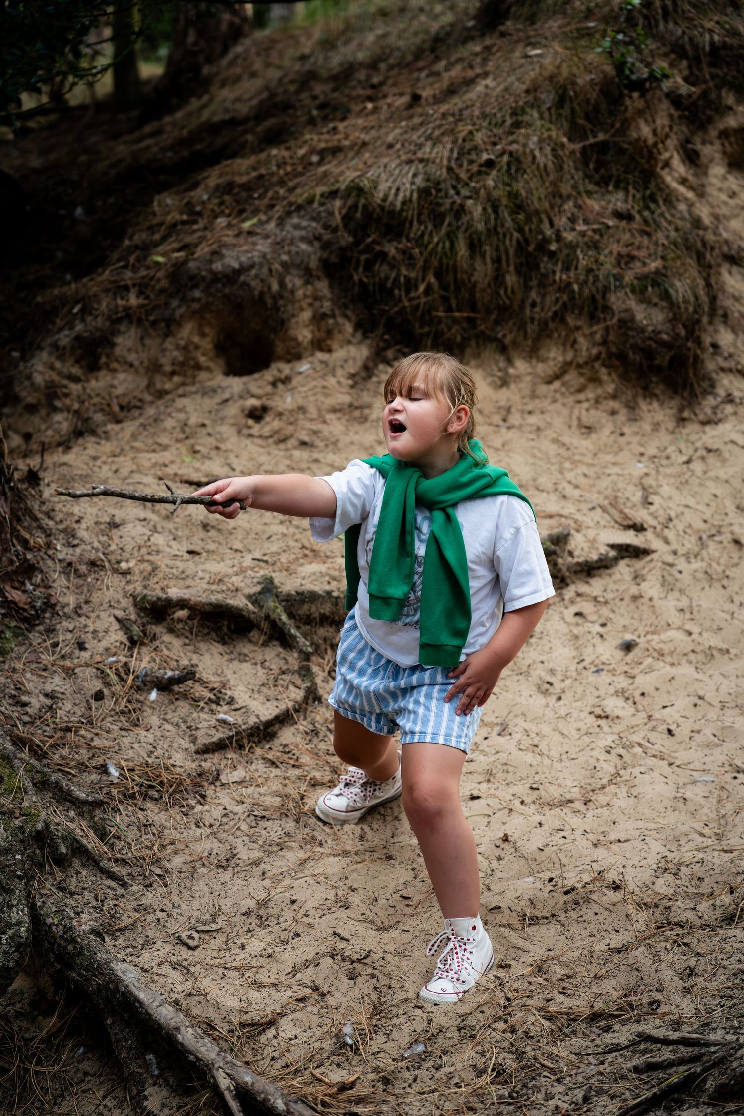 Young girl in green scarf and blue shorts pointing a stick while exploring a sandy woodland path.