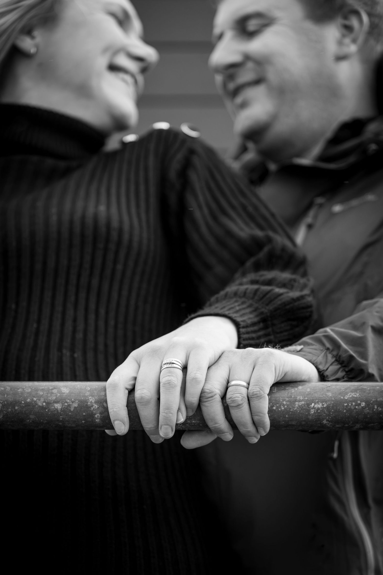 Black and white photo of a married couple smiling at each other, their hands with wedding rings resting on a railing.