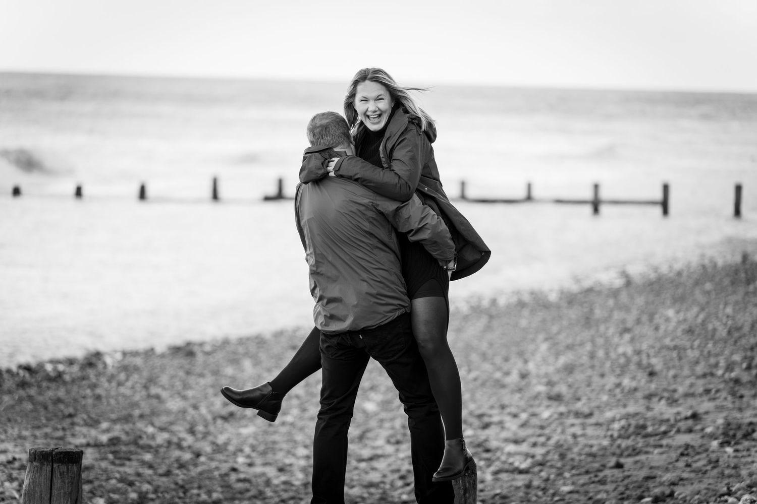 Black and white photo of a man lifting a laughing woman in the air on a pebble beach with wooden groynes and the sea behind.