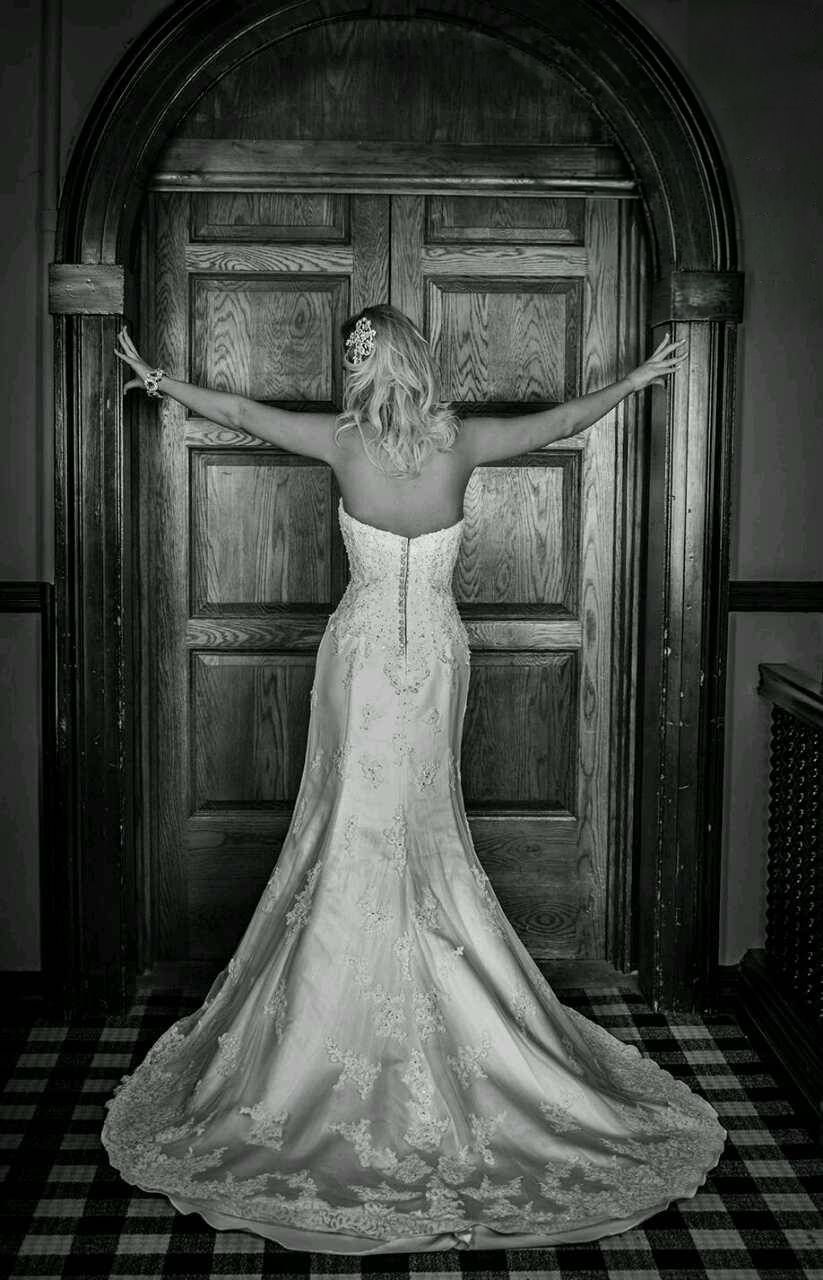 Black and white photo of a bride in a lace wedding gown with a long train, arms outstretched against a tall paneled wooden door.