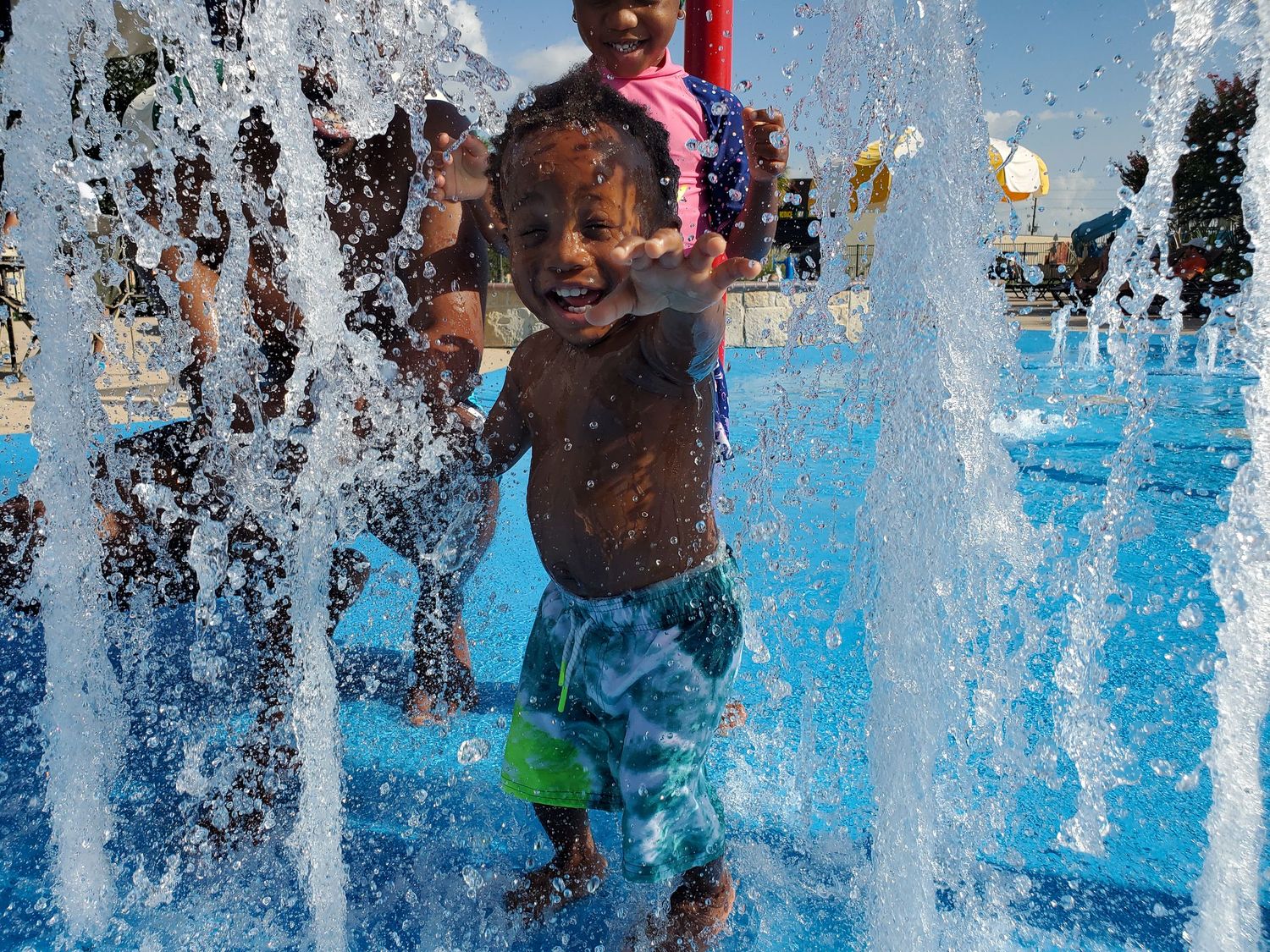 A young boy at a splash park reaches joyfully for the camera as water rises up around him.