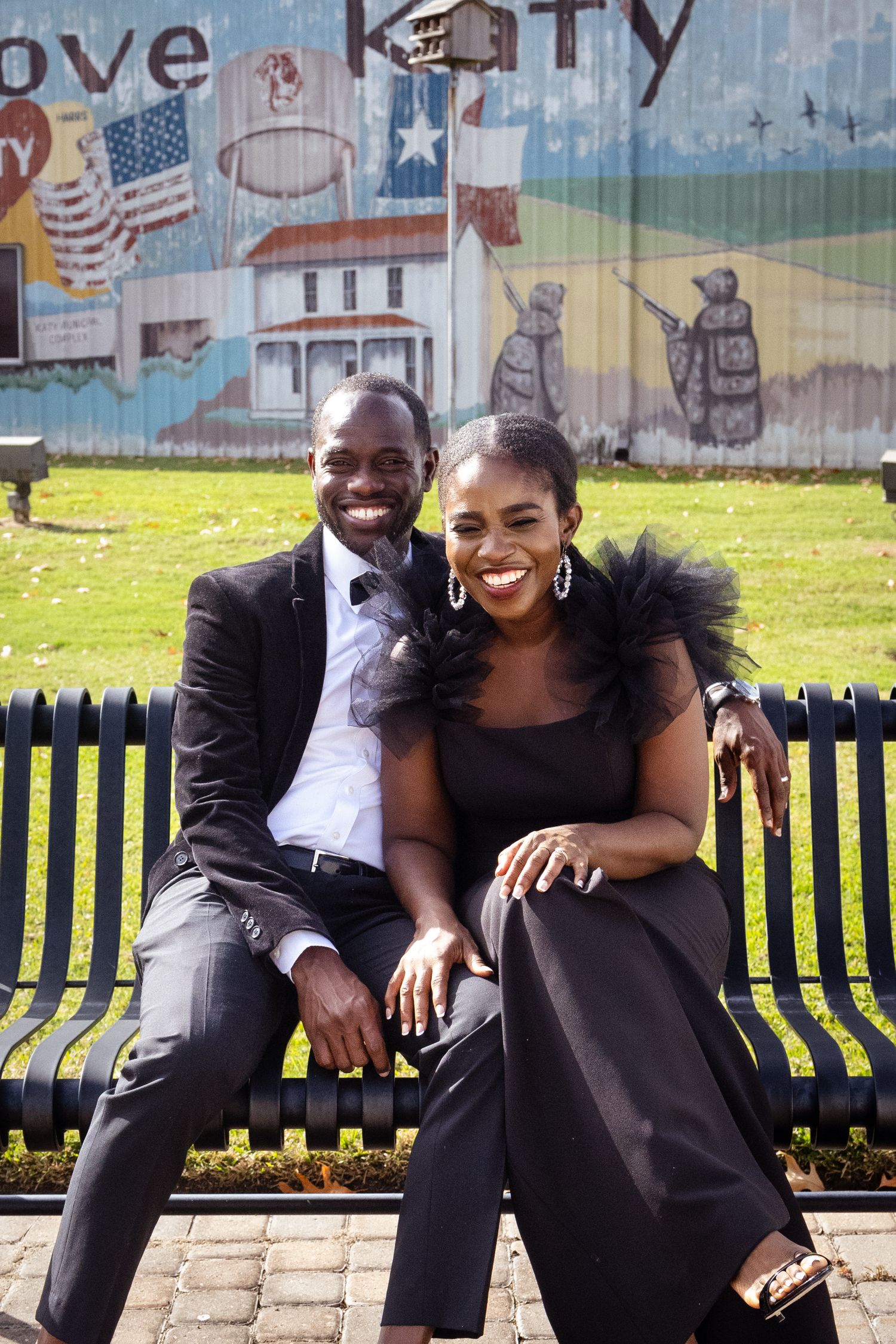 A couple dressed in evening wear laughs together on a park bench.
