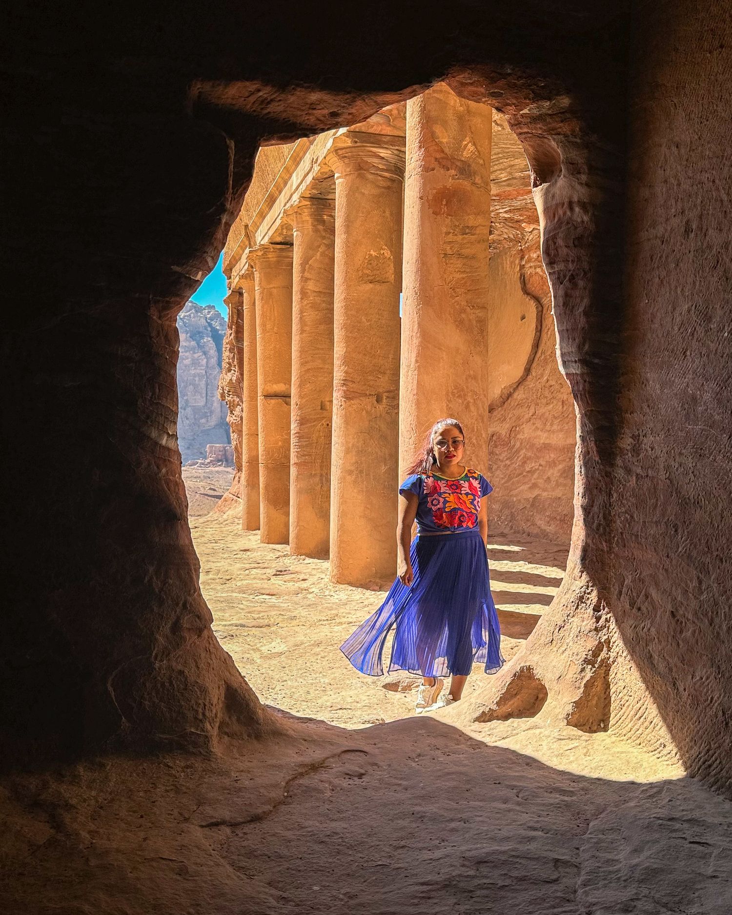 Woman in a blue dress walks through the opening of a cave in the desert.