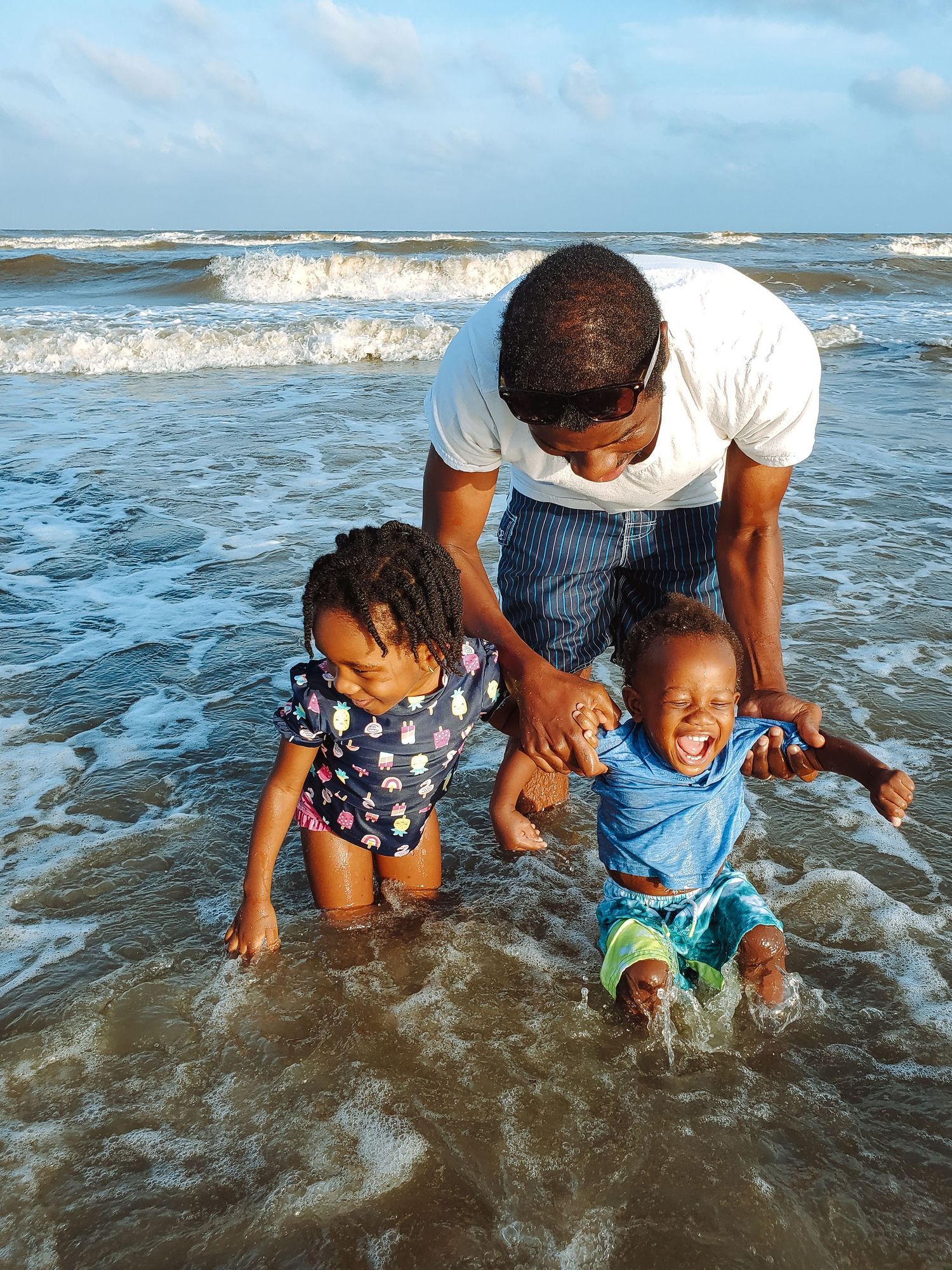 A father holds his laughing son and daughter at the beach while gentle waves approach them.