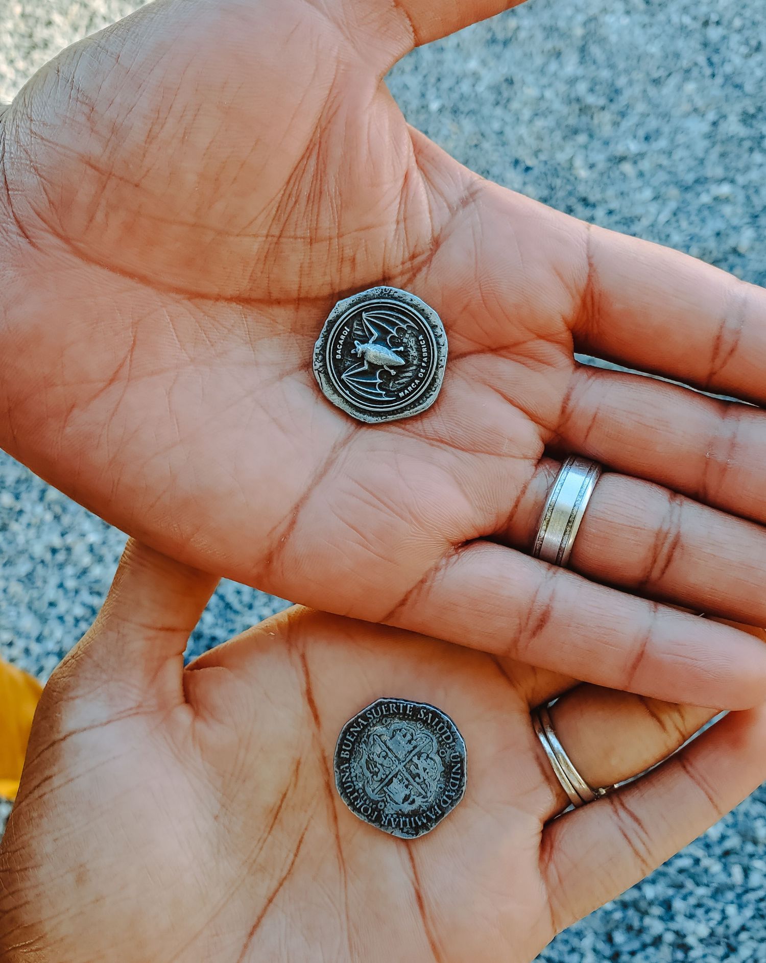 Two hands with wedding bands hold up silver tokens that grant entry into a rum factory.