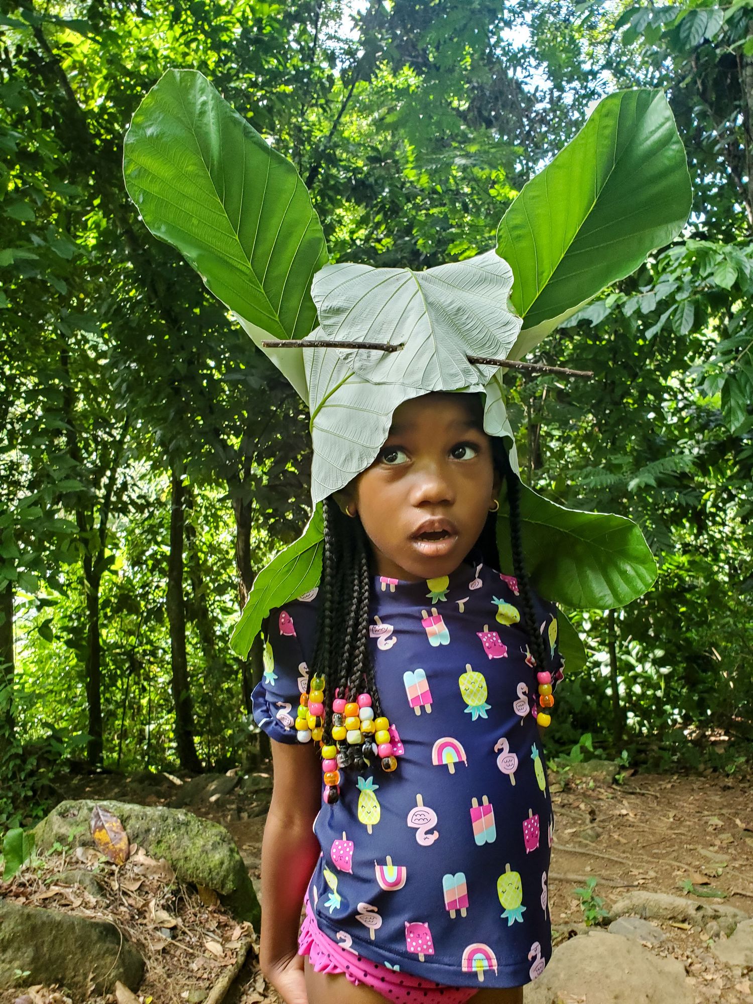 A young girl by the water in the jungle wears a large hat made of leaves and twigs.