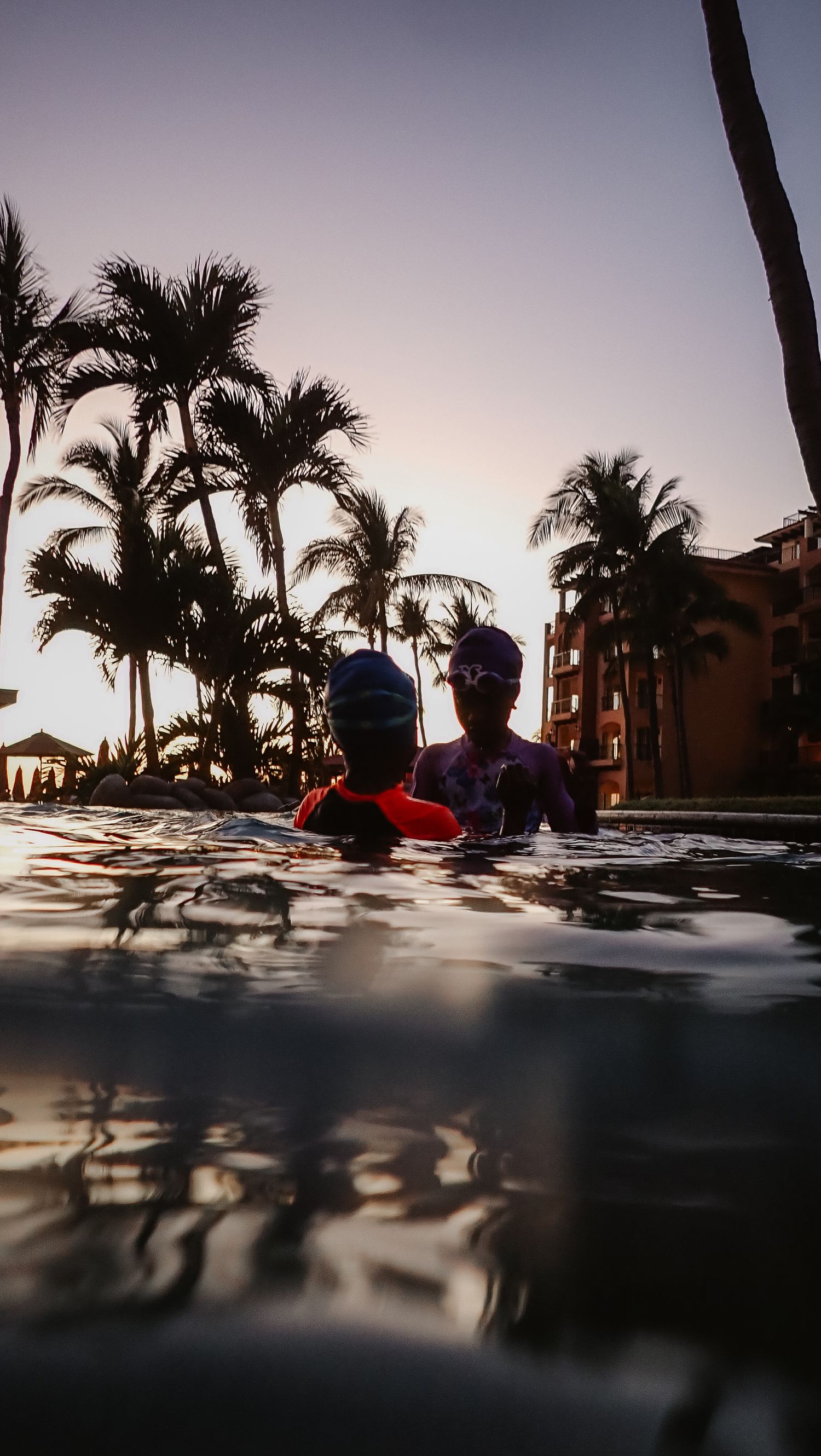 Two children playing in a pool, silhouetted against a palm trees at sunset.