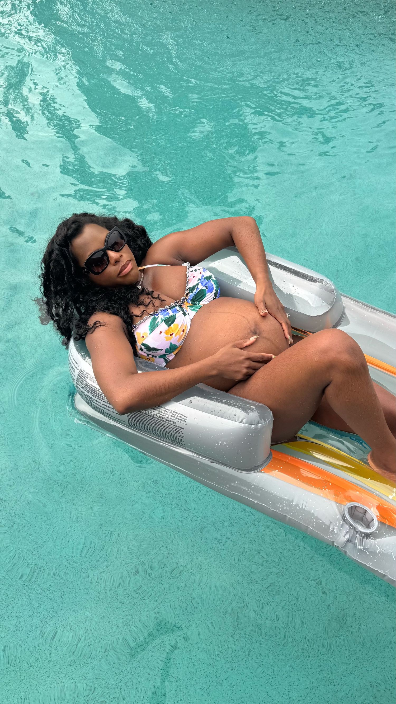 A pregnant woman holds her belly while relaxing on a colorful floating bed in a pool.