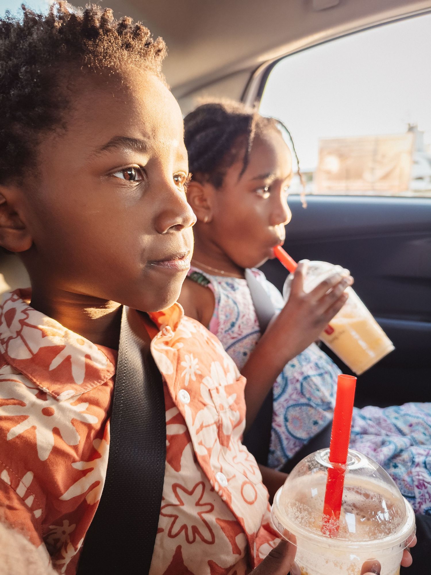 Two tired children sit in the back seat of a car, drinking milkshakes.