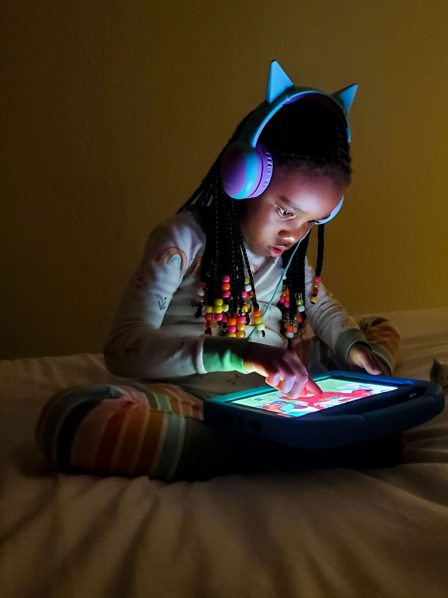 A young girl sits on her bed with cat-ear headphones, bathed in the light of her tablet device.