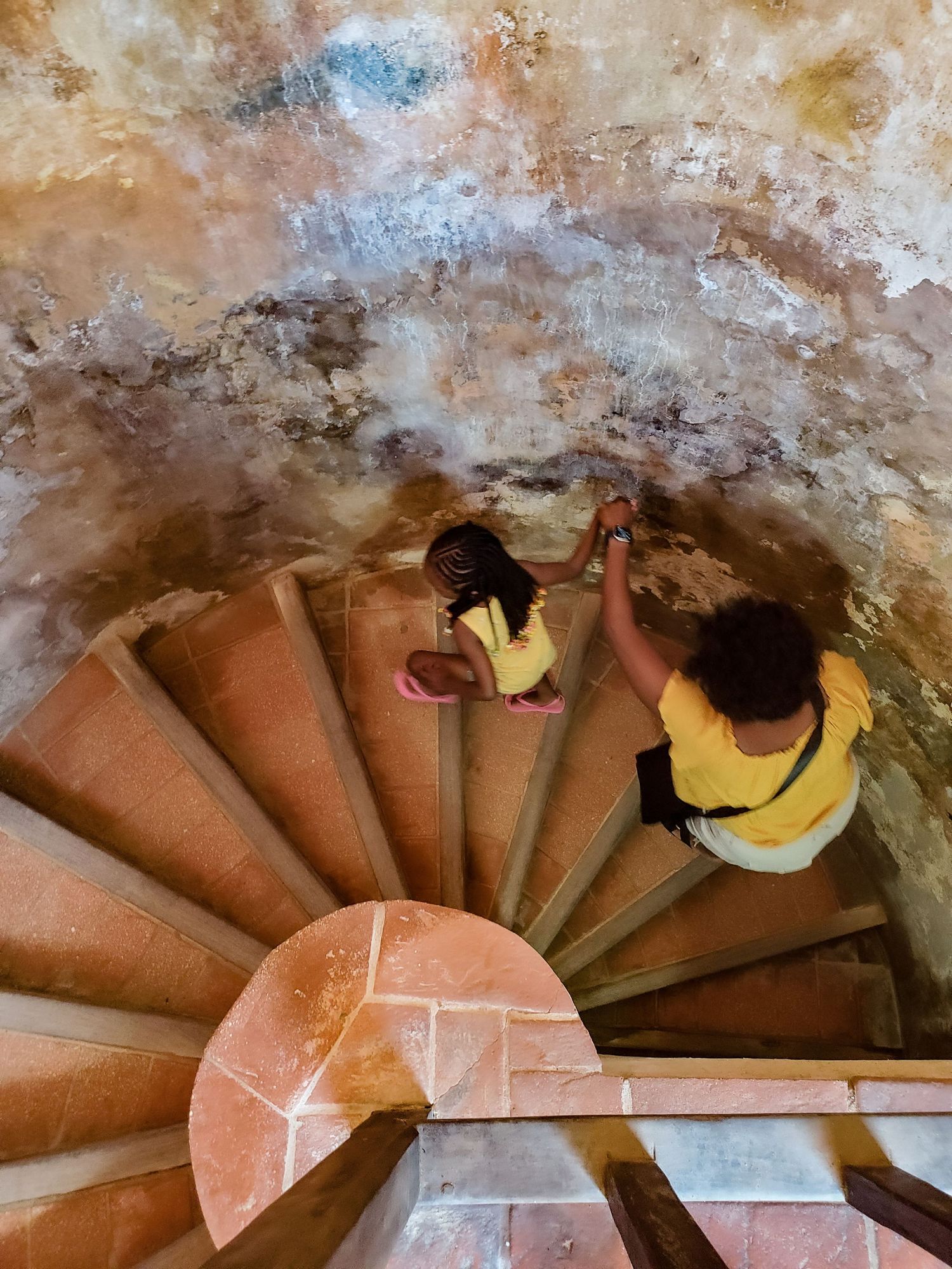 A grandmother and granddaughter, both wearing yellow, climb up terracotta colored spiral stairs.