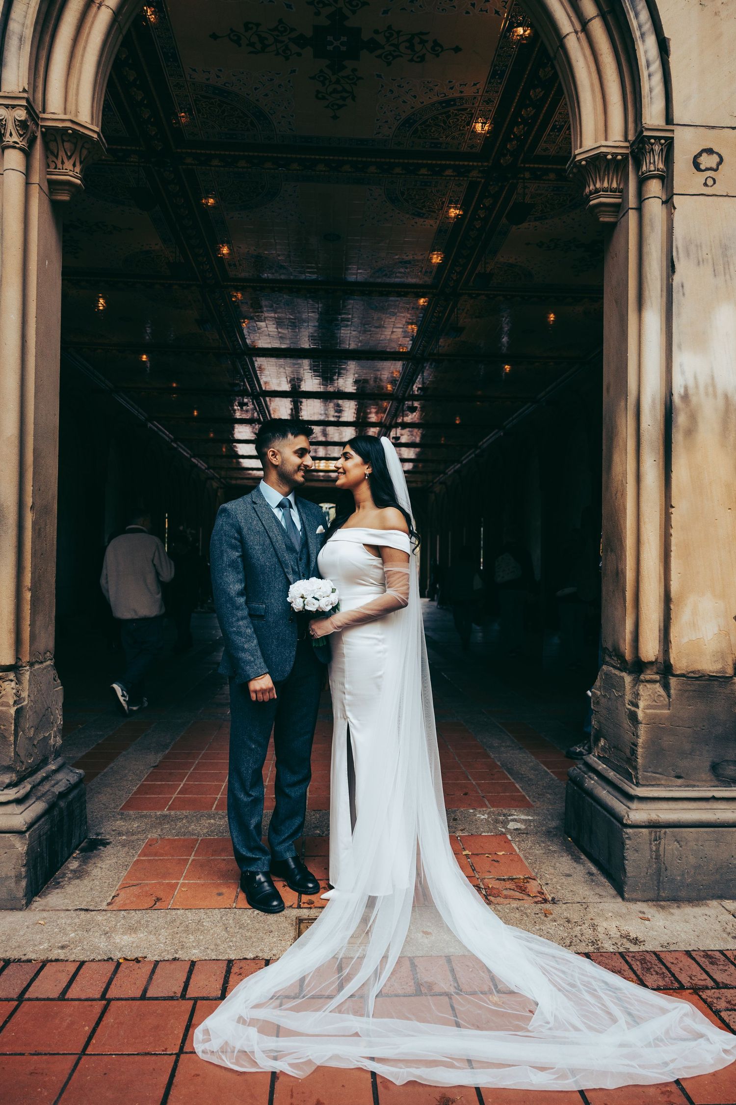 Bride and groom pose under ornate stone arch, bride in elegant white gown with long veil holding bouquet.