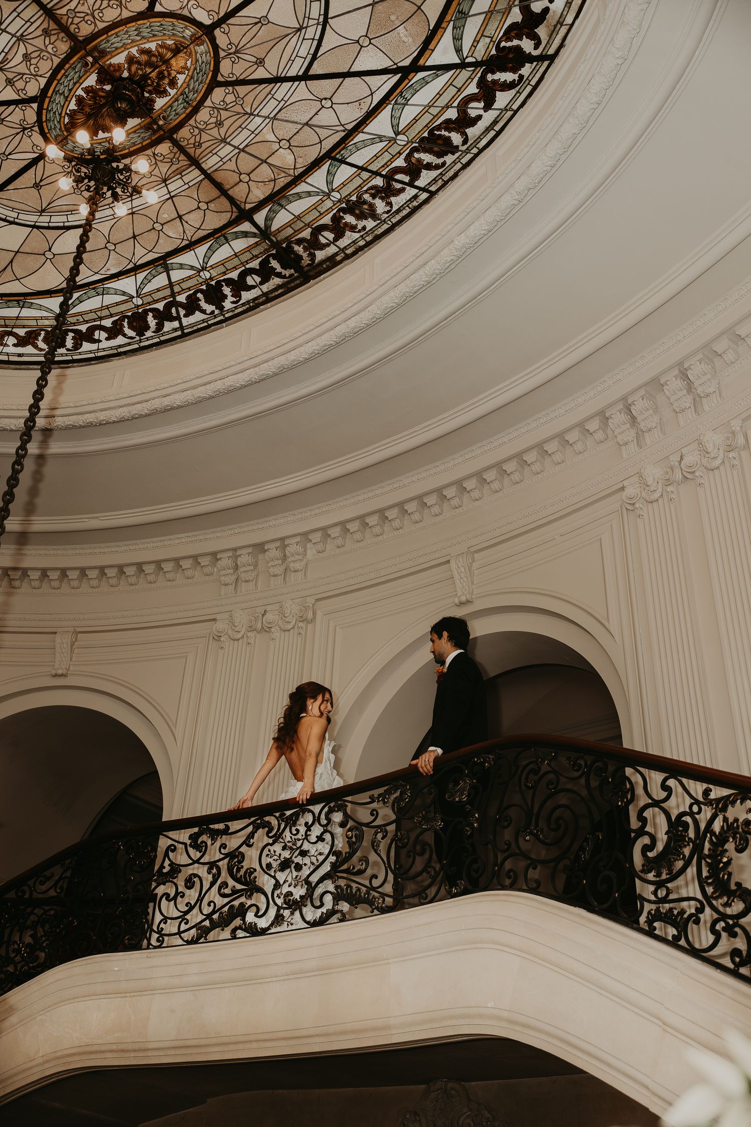 Bride and groom stand on ornate iron balcony beneath stained glass dome in elegant white wedding venue.