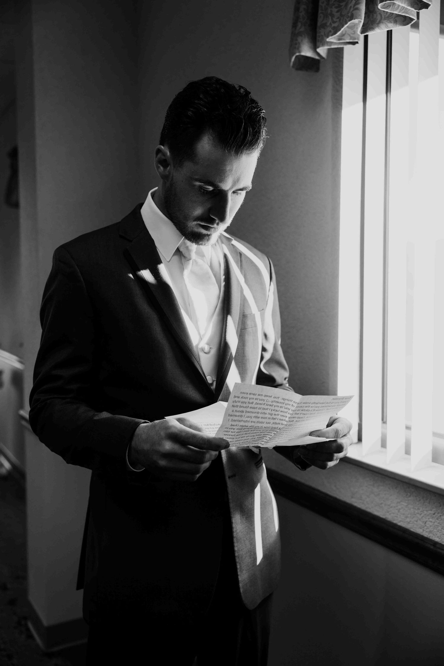 Black and white photo of a groom in a suit reading a heartfelt letter by a window on his wedding day.