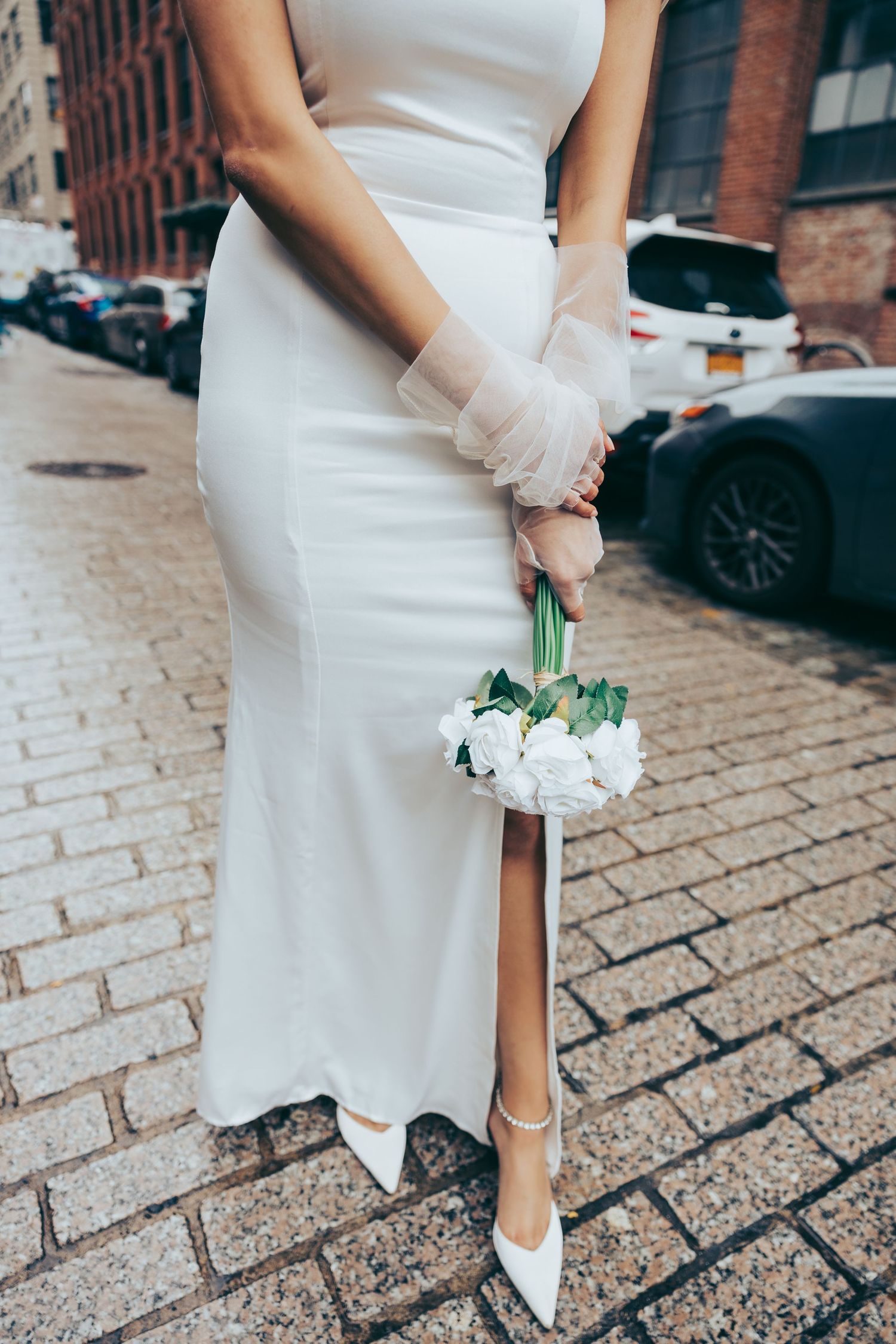 Bride in white gown holding small white rose bouquet, standing on cobblestone street in white heels.