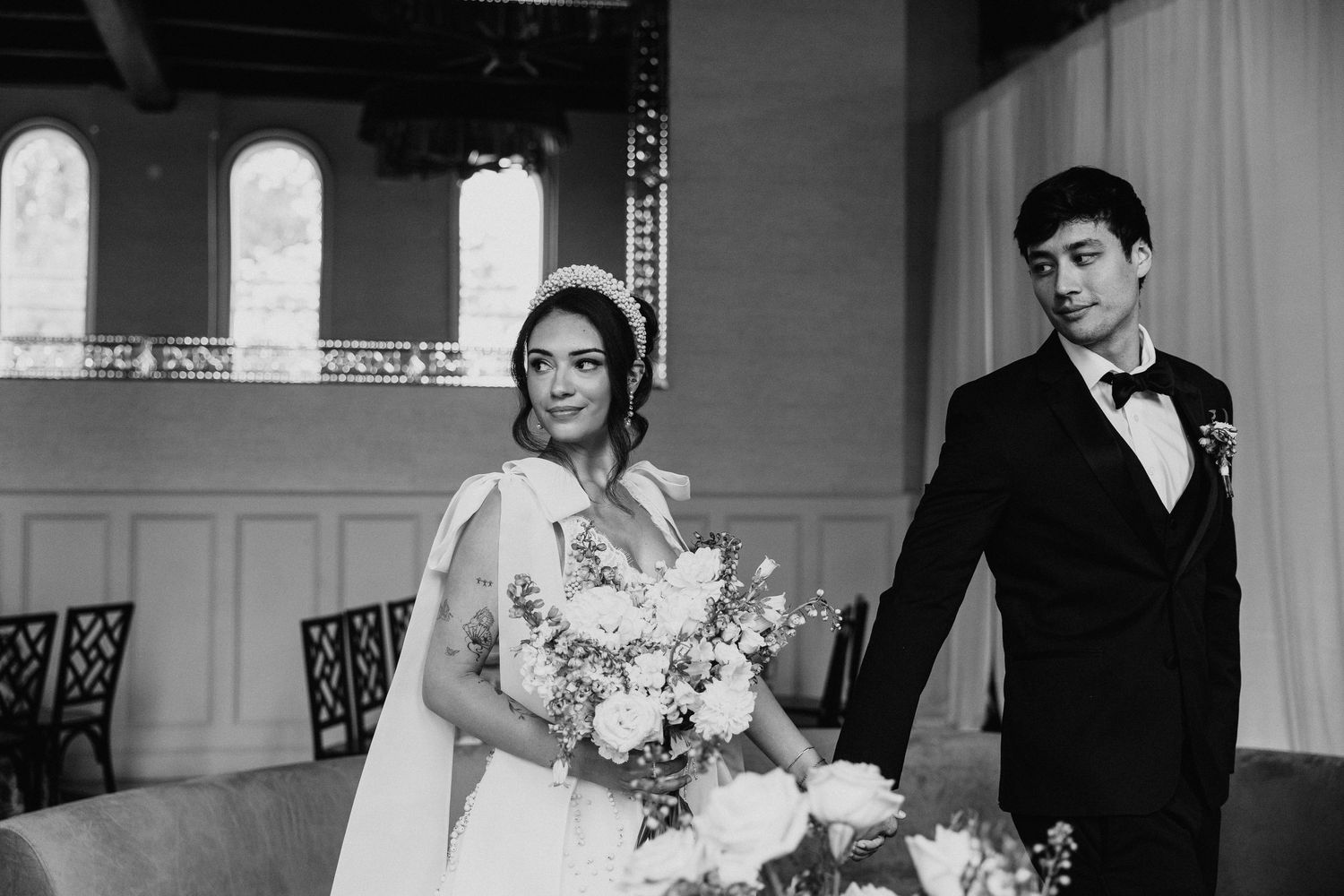 Black and white photo of a bride holding a bouquet and groom in tuxedo standing in an elegant wedding venue. Elkins estate wedding venue.
