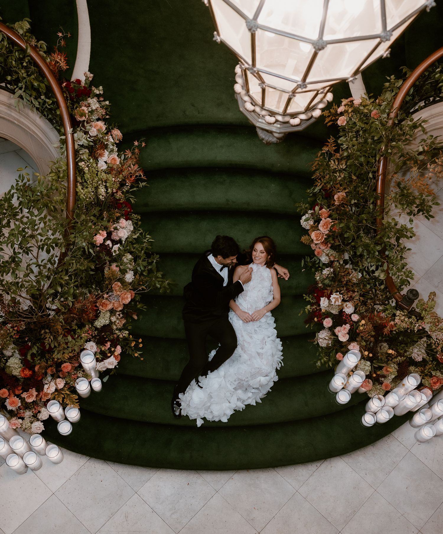 Bride and groom embracing on a green velvet staircase surrounded by lush floral arrangements and candles.