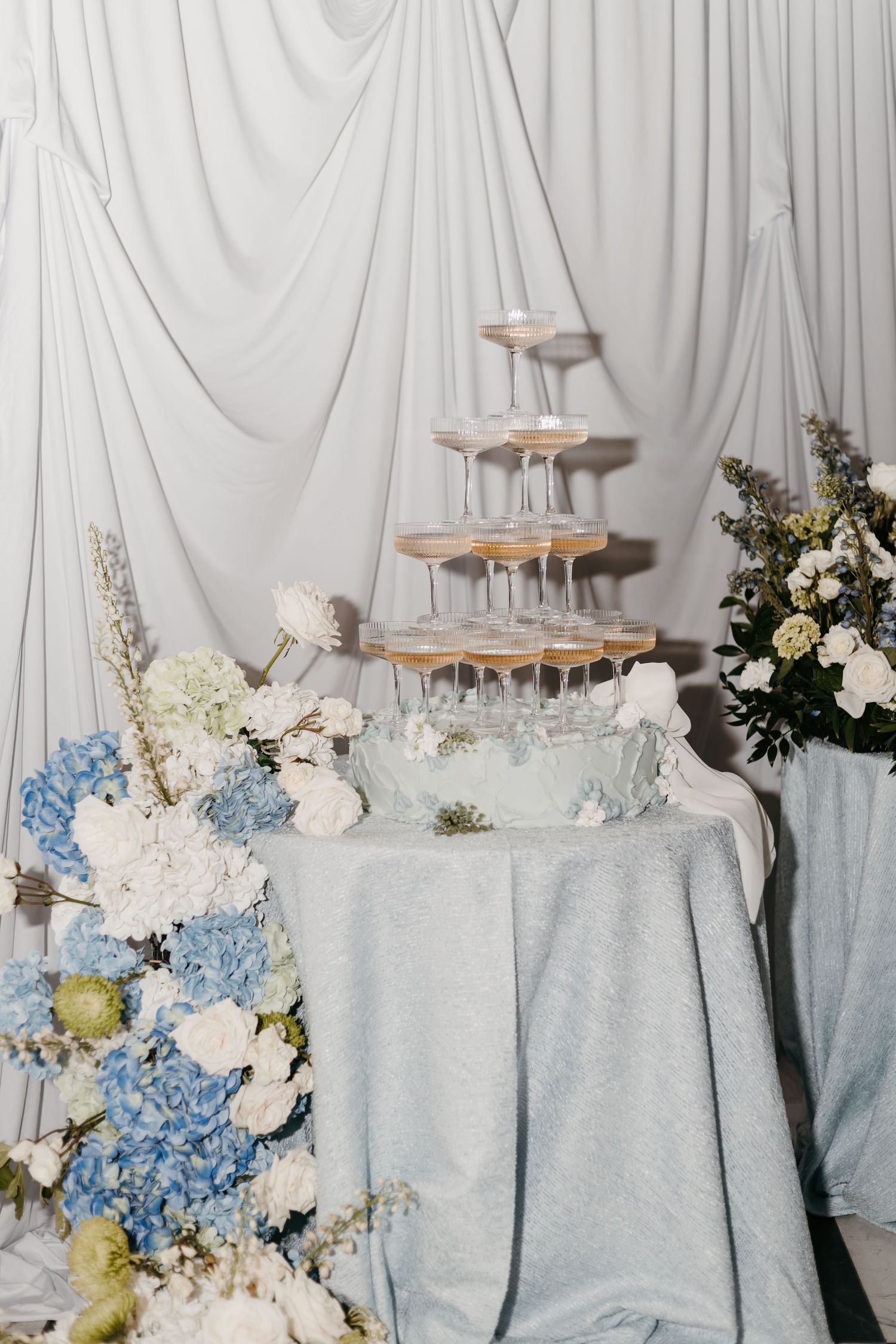 Elegant wedding champagne tower with blue hydrangeas and white flowers on a light blue draped table.