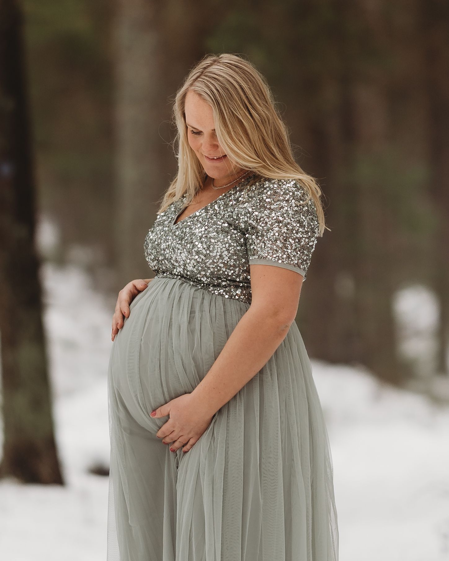 Gravid kvinna i glittrande klänning stående i snö i skogen