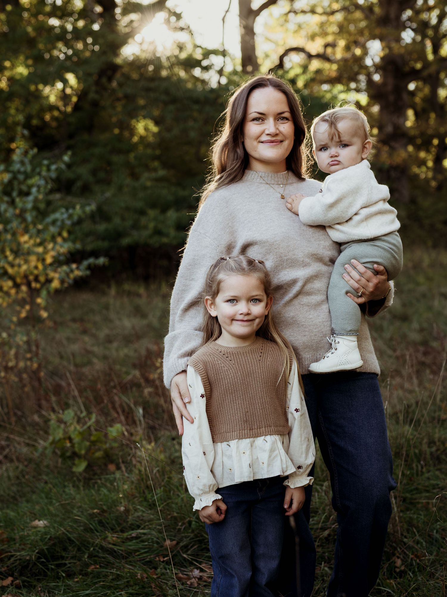 Mamma med två flickor ståendes bland träden i vårsolen.
