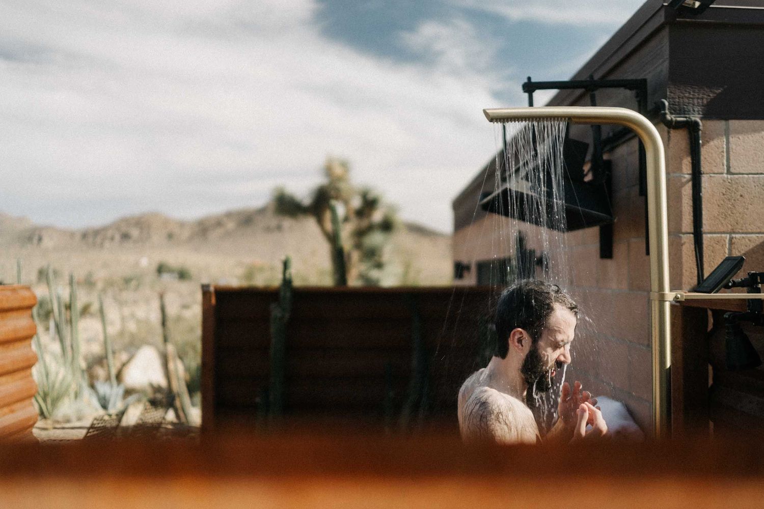 Bearded man at an outdoor shower beside a desert house, Joshua trees and hills behind