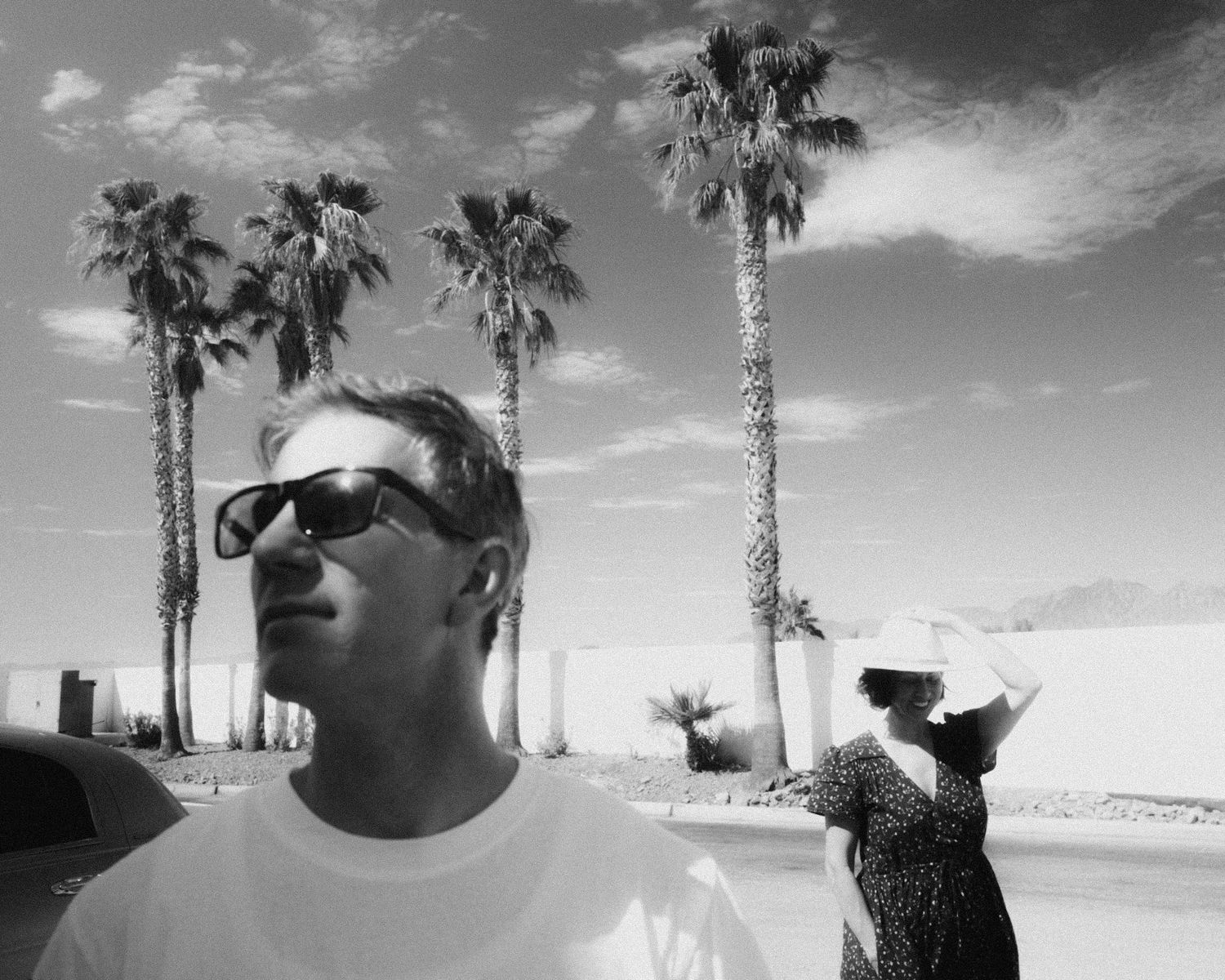 Black and white photo of a man in sunglasses and a white T-shirt with palm trees behind him and a woman in a hat in the background.