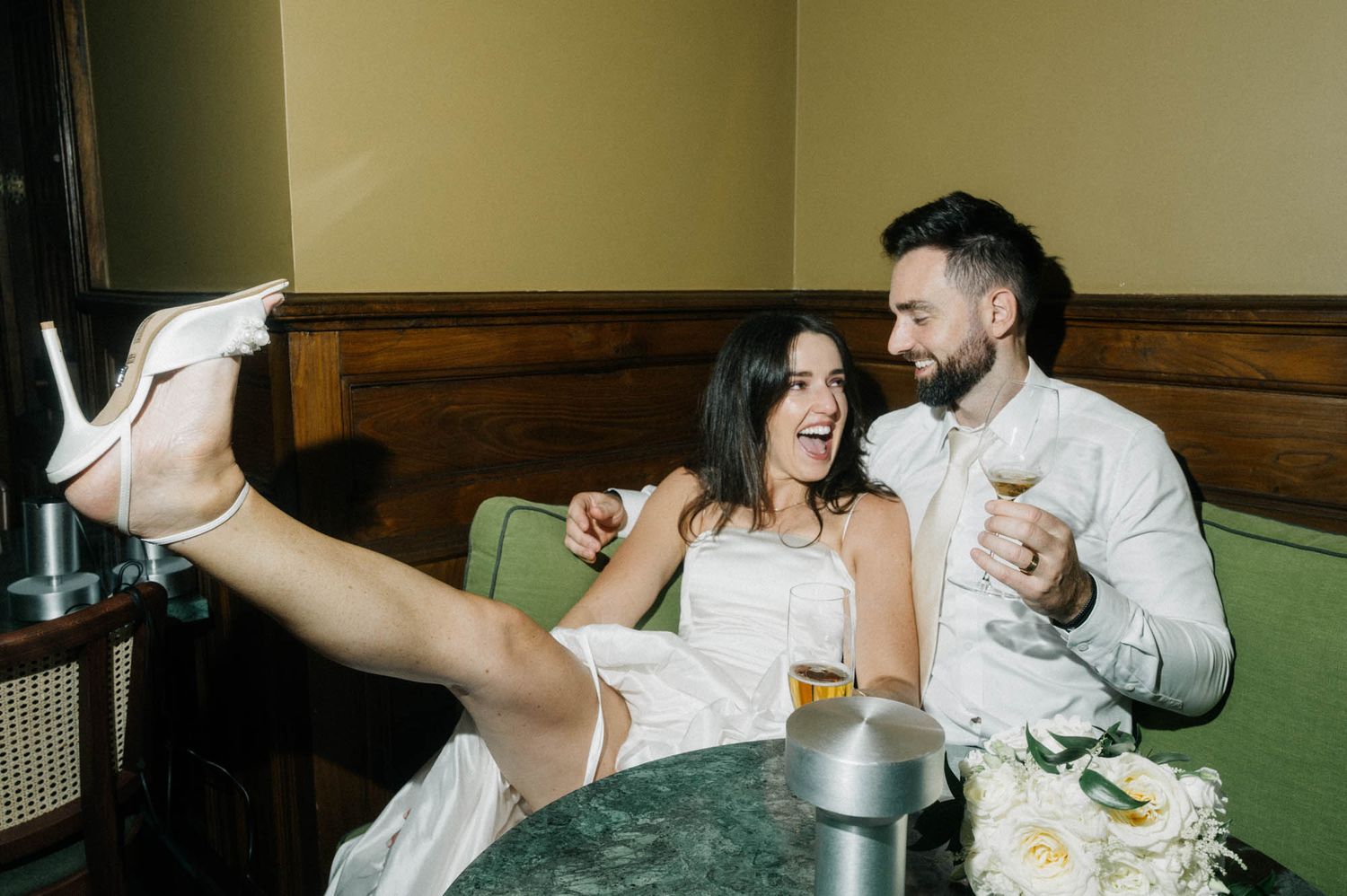 Couple laughing on a green sofa in a bar, the woman in a white dress with one leg raised.