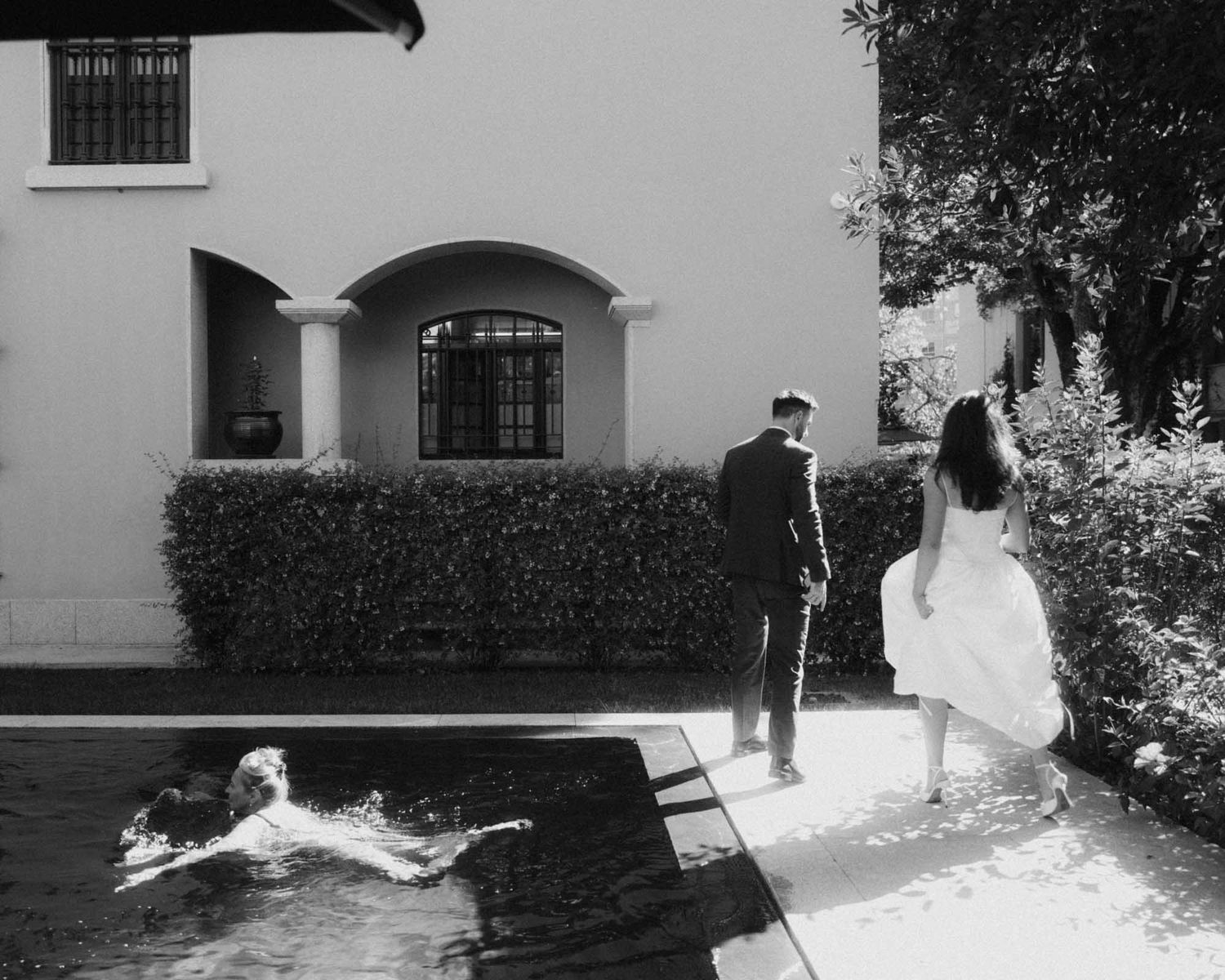 Black and white photo of a man and a woman in a white dress walking along a garden path beside a pool, with a swimmer in the water.