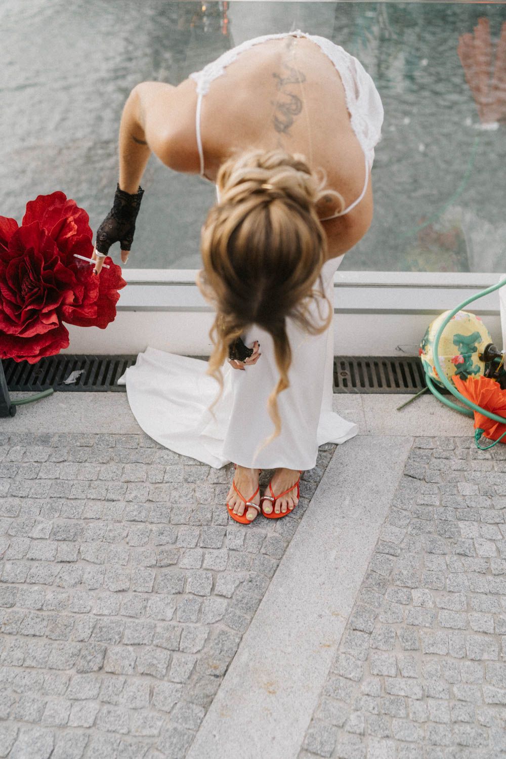 Barefoot bride in a white dress bending over to look down from a window, red flowers beside her