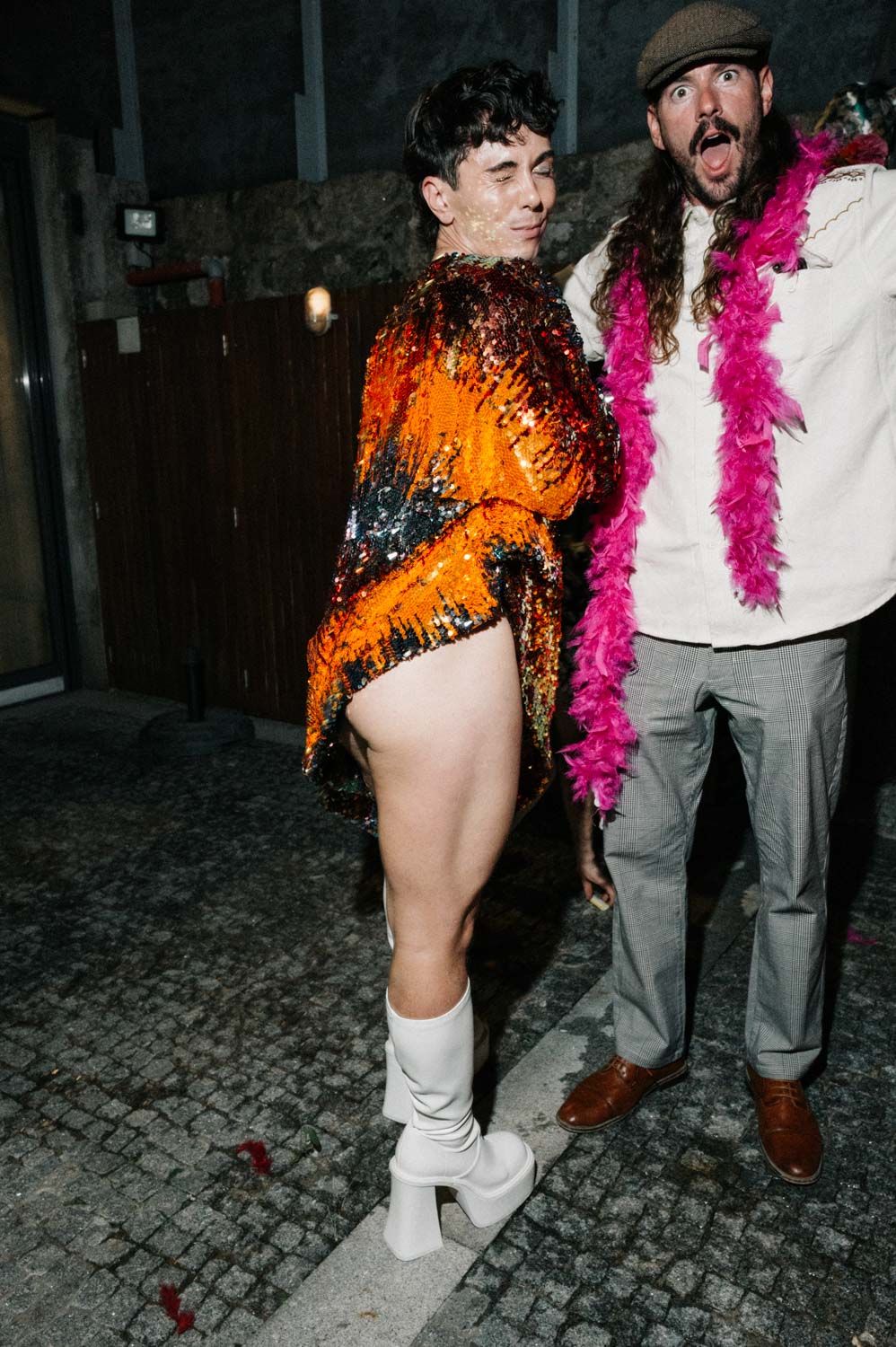 Two people on a cobbled street at night: one in an orange sequin dress and white platform boots, the other in a flat cap and white shirt wea