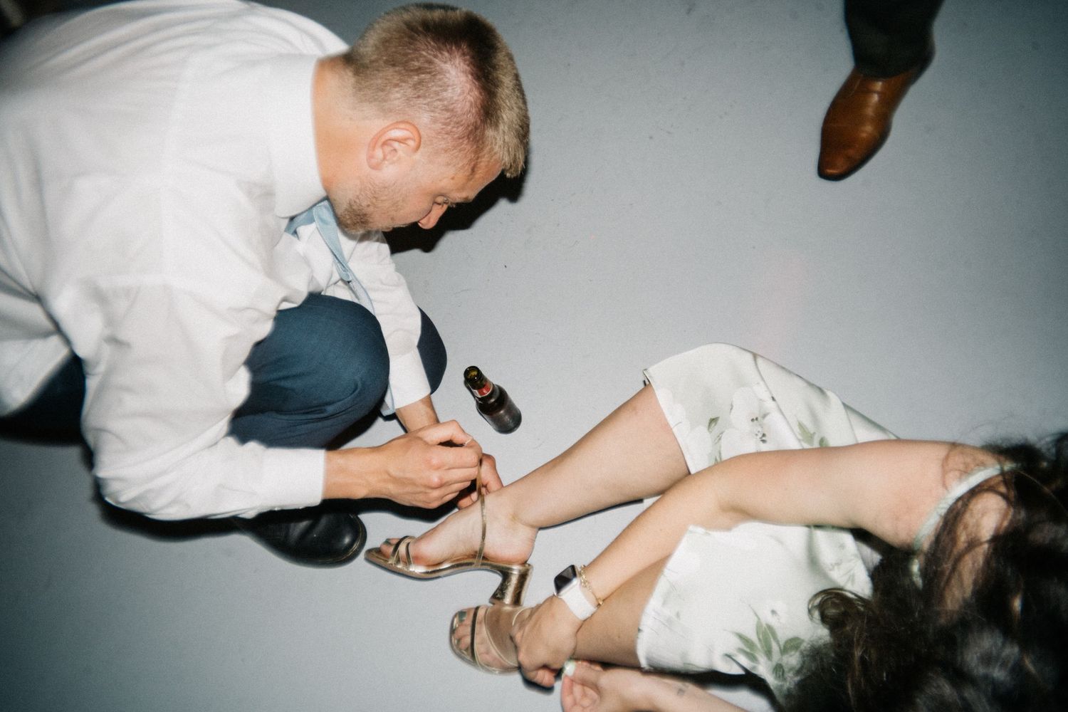 Wedding guest kneeling to help fasten a woman's sandal in a candid, flash-lit moment.