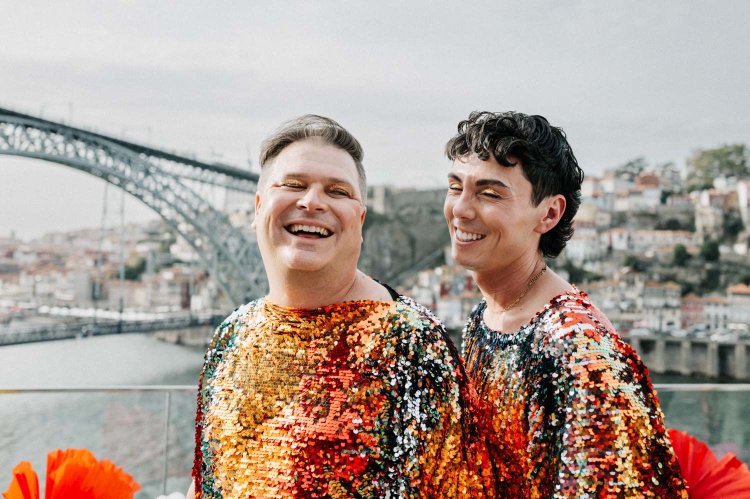 Two grooms laughing in sequin outfits with the Porto bridge behind them, same-sex wedding photography in Porto, Portugal