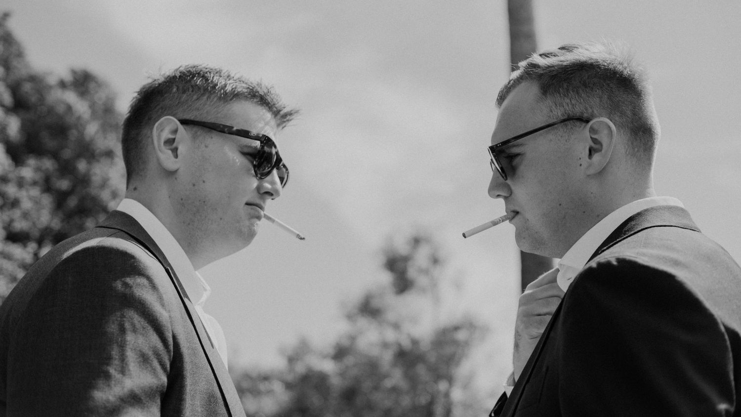 Black and white photo of two men in suits and sunglasses, each with a cigarette in his mouth, facing each other.