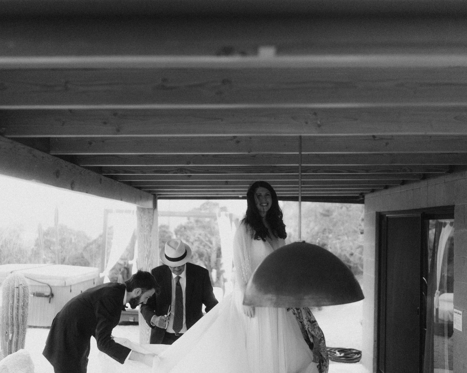 Black and white photo of a bride on a porch while two men adjust her gown