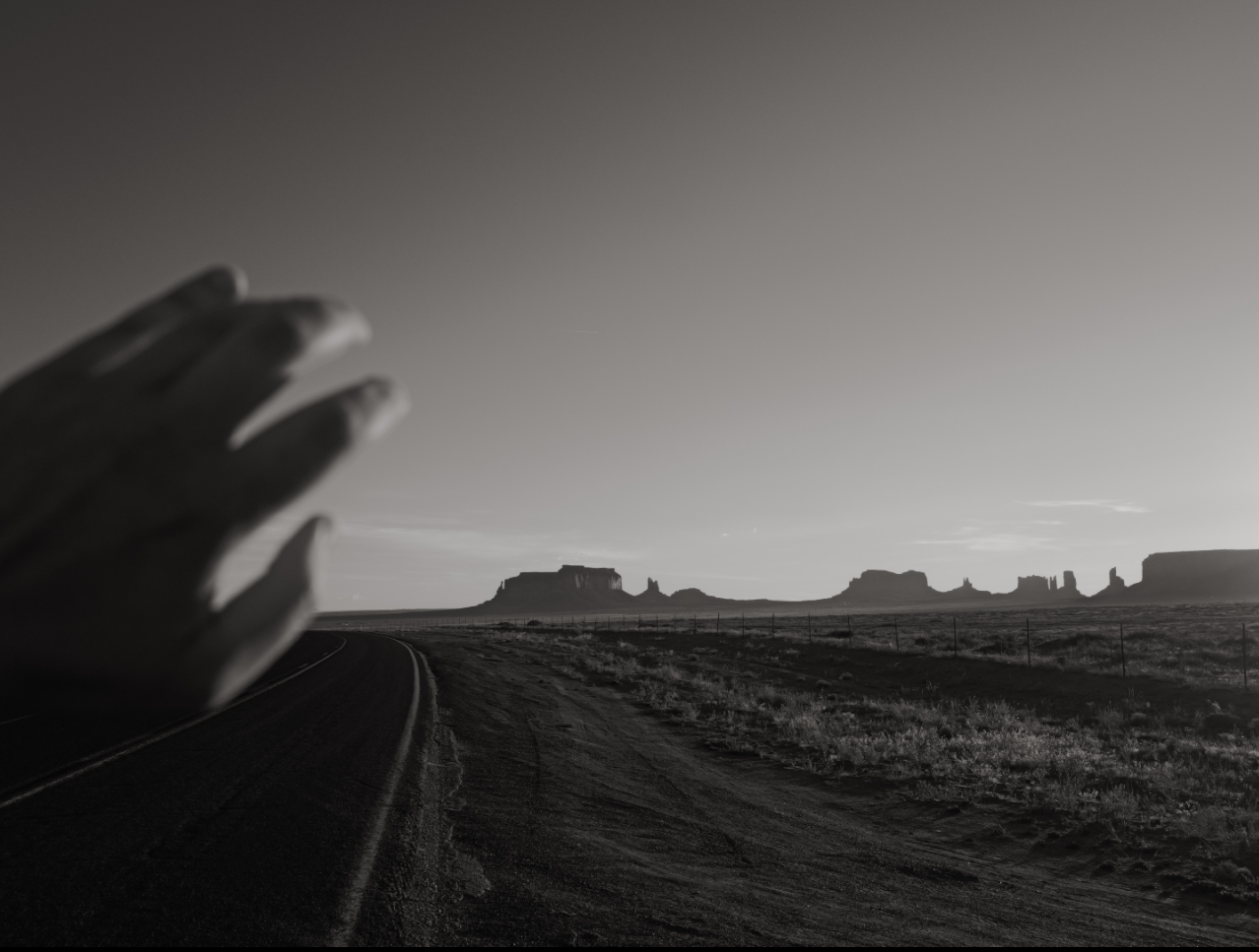 Black and white view of a desert road and distant rock formations, with a hand raised in the foreground.