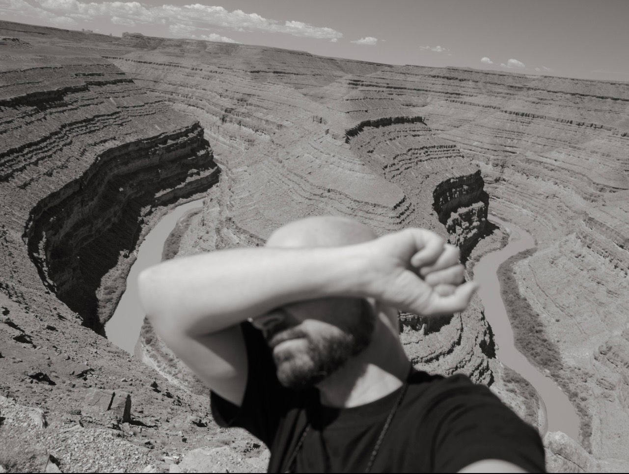 Man in a black T-shirt covering his face with his arm above a winding canyon.