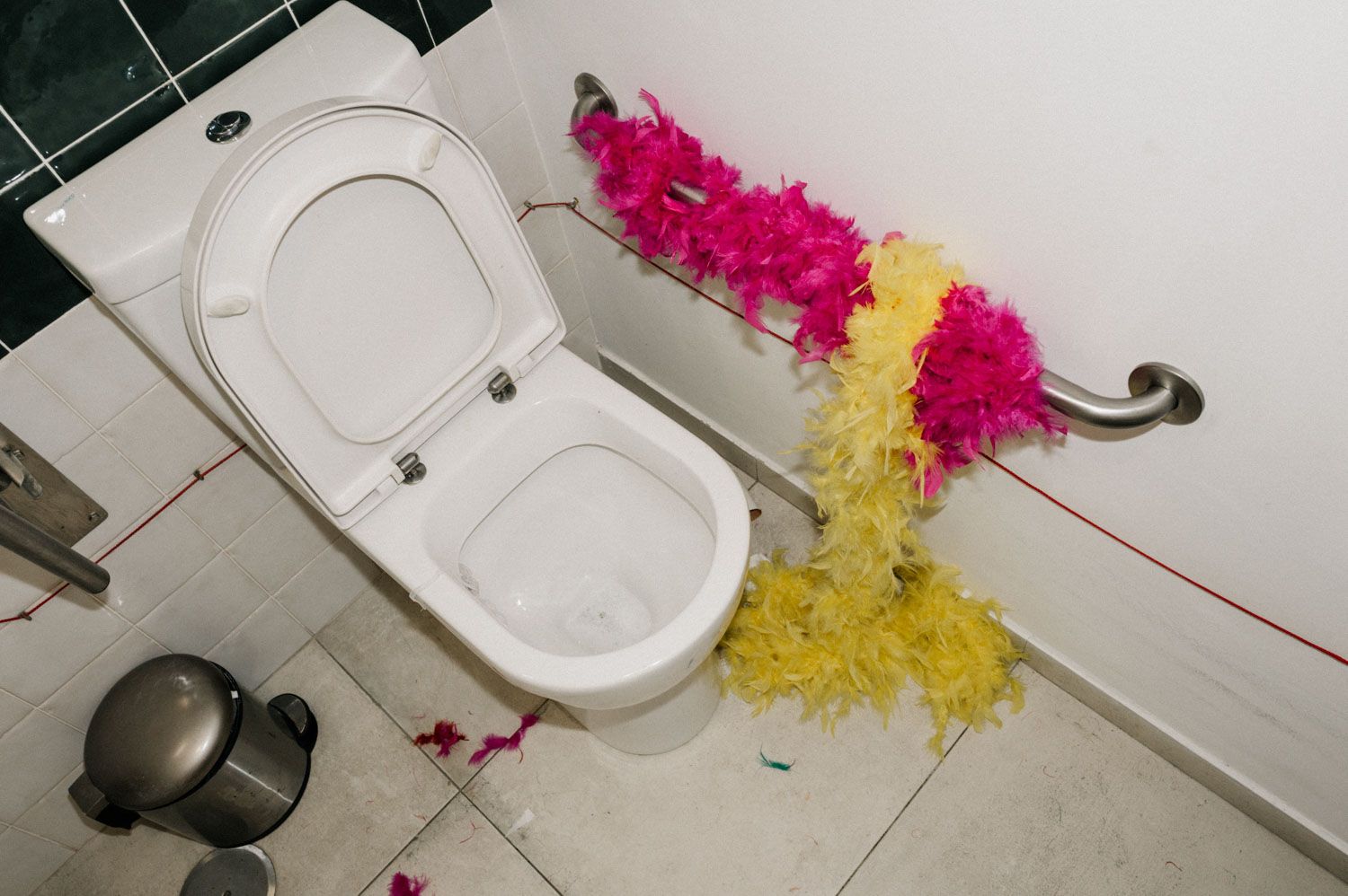 Pink and yellow feather boas draped over a grab rail and the floor beside a toilet.