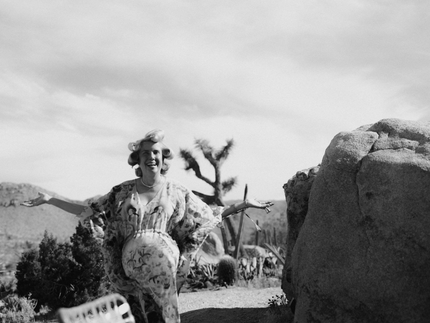 Black and white photo of a laughing woman in a patterned dress, arms open beside a boulder and Joshua tree