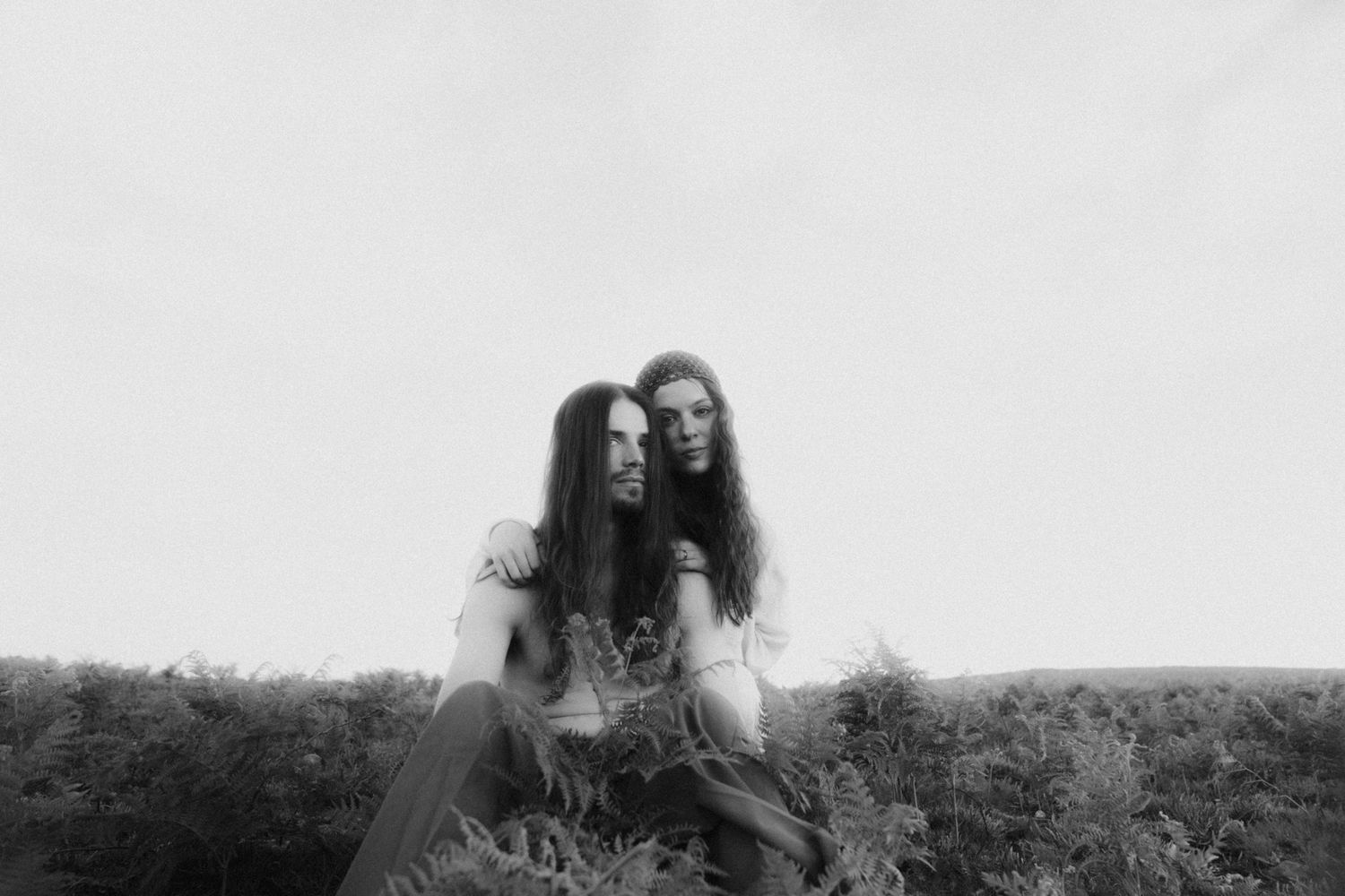 Black and white portrait of a couple with long hair sitting close together among ferns.