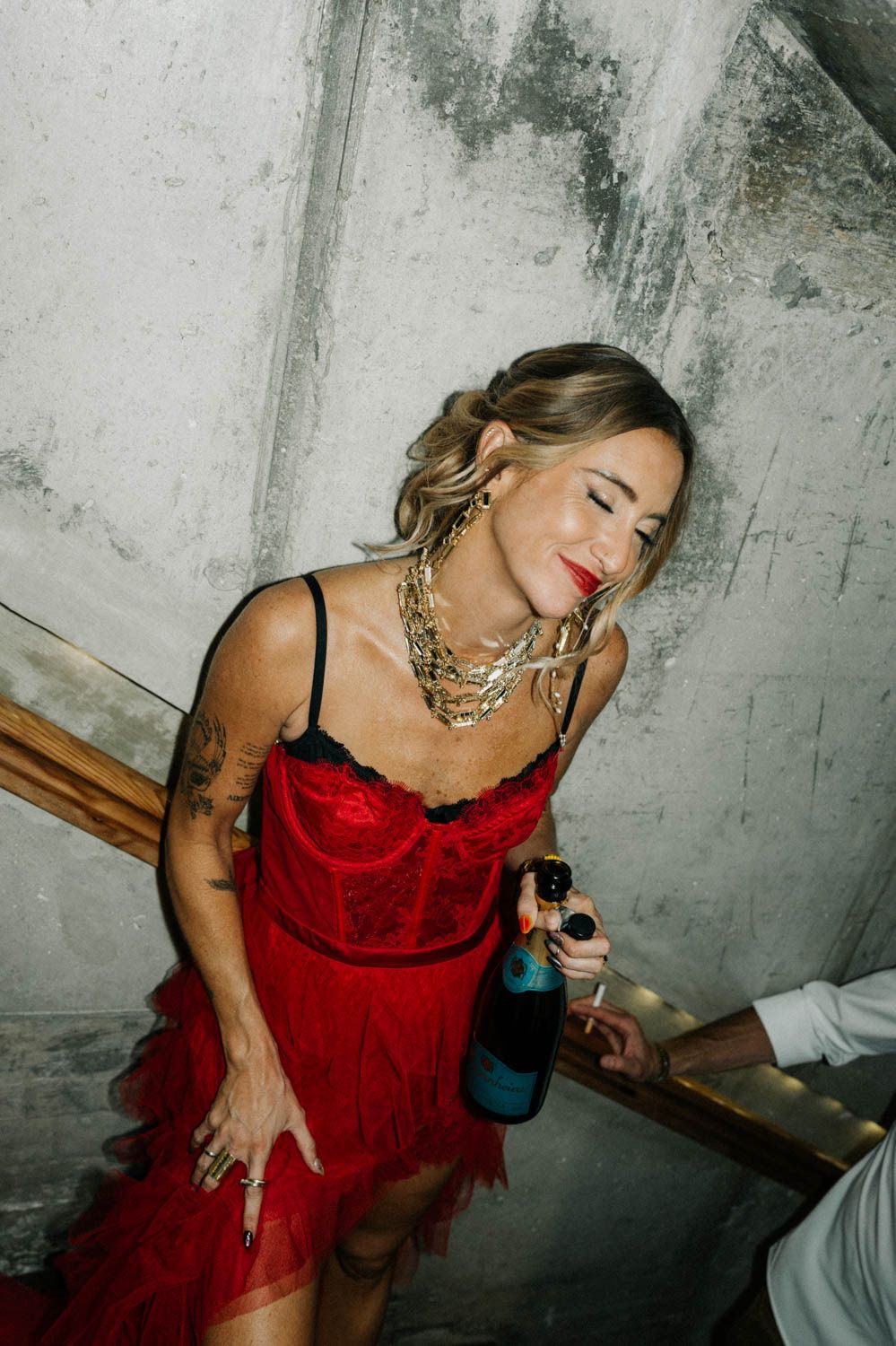 Woman in a red dress and gold necklaces holding a bottle, standing against a concrete wall beside a stair railing.