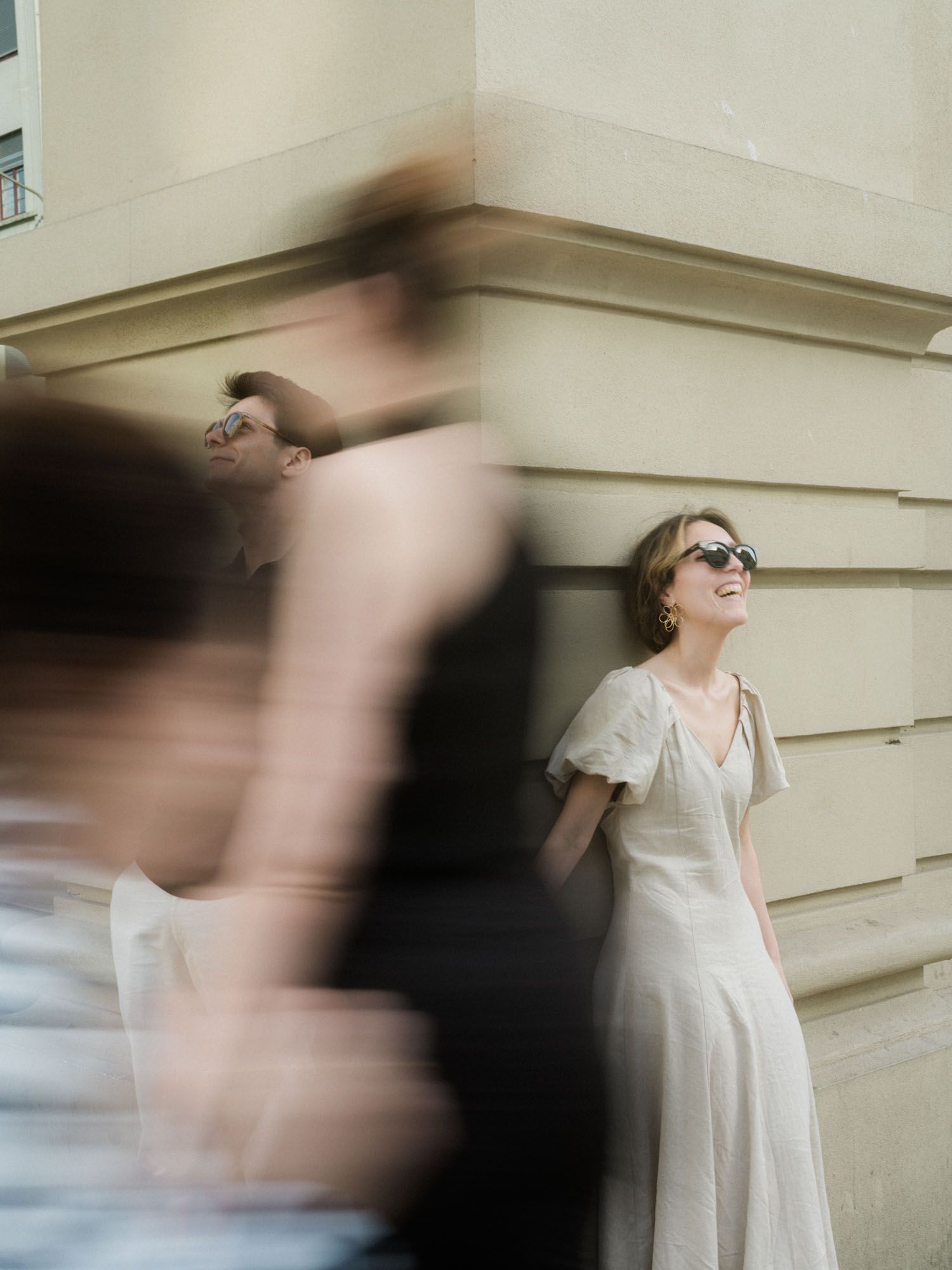 Woman in a cream dress leaning against a stone wall while two blurred people walk past.