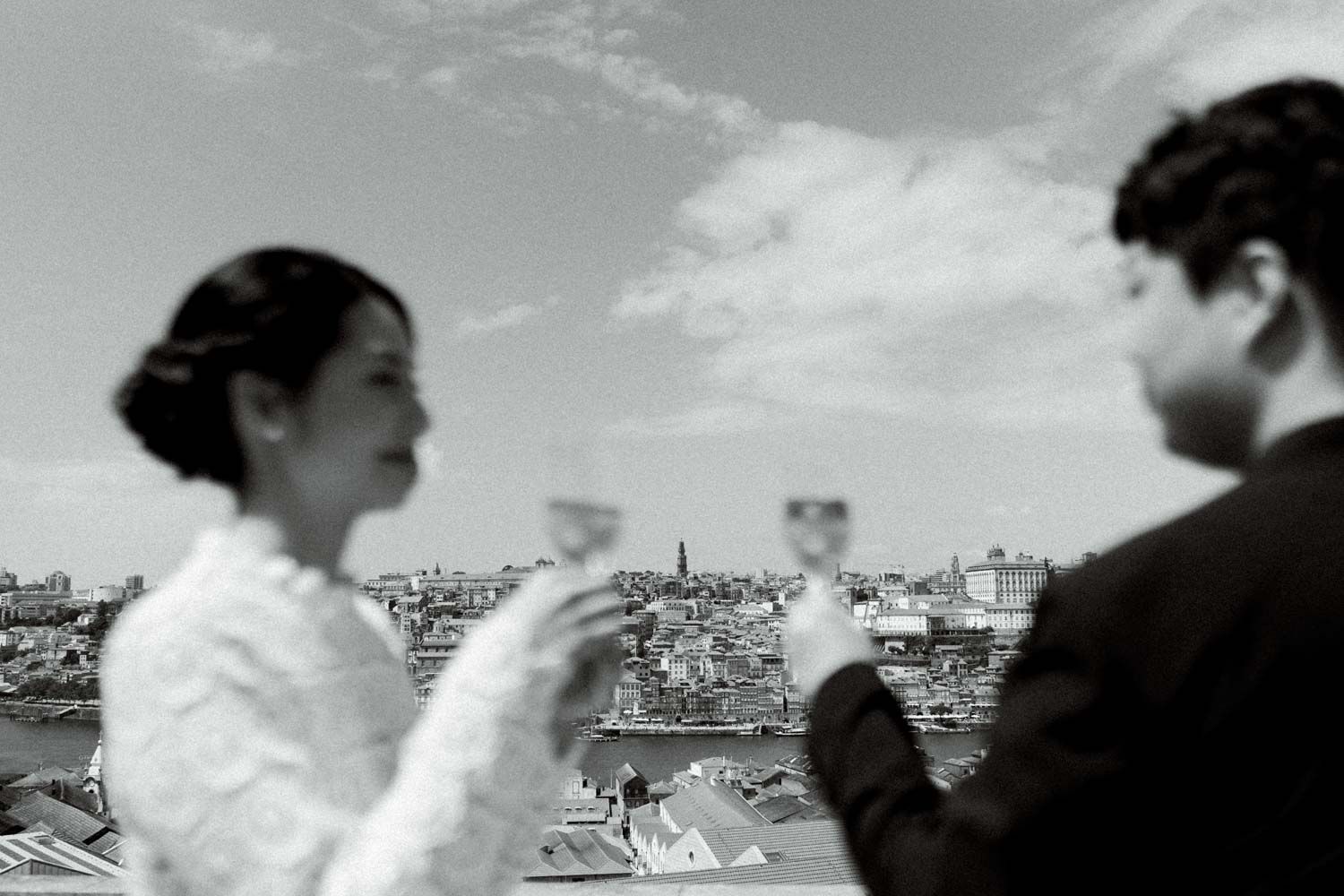 Black and white photo of a couple raising their glasses in a toast with a city view behind them