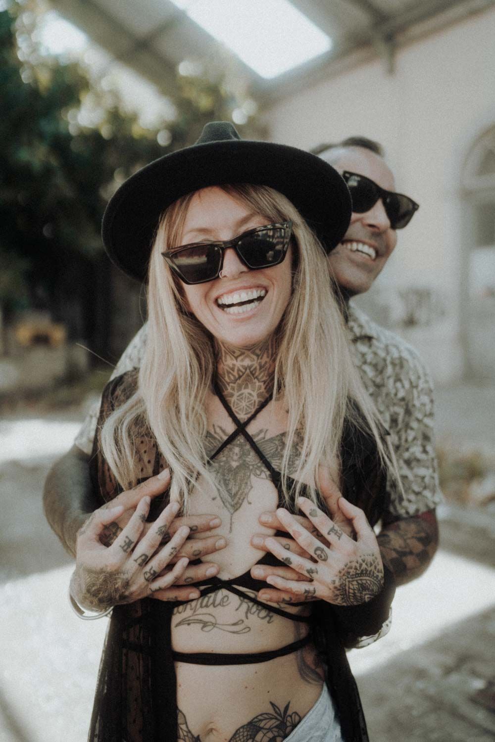 Woman in a black hat and sunglasses laughing as a man hugs her from behind