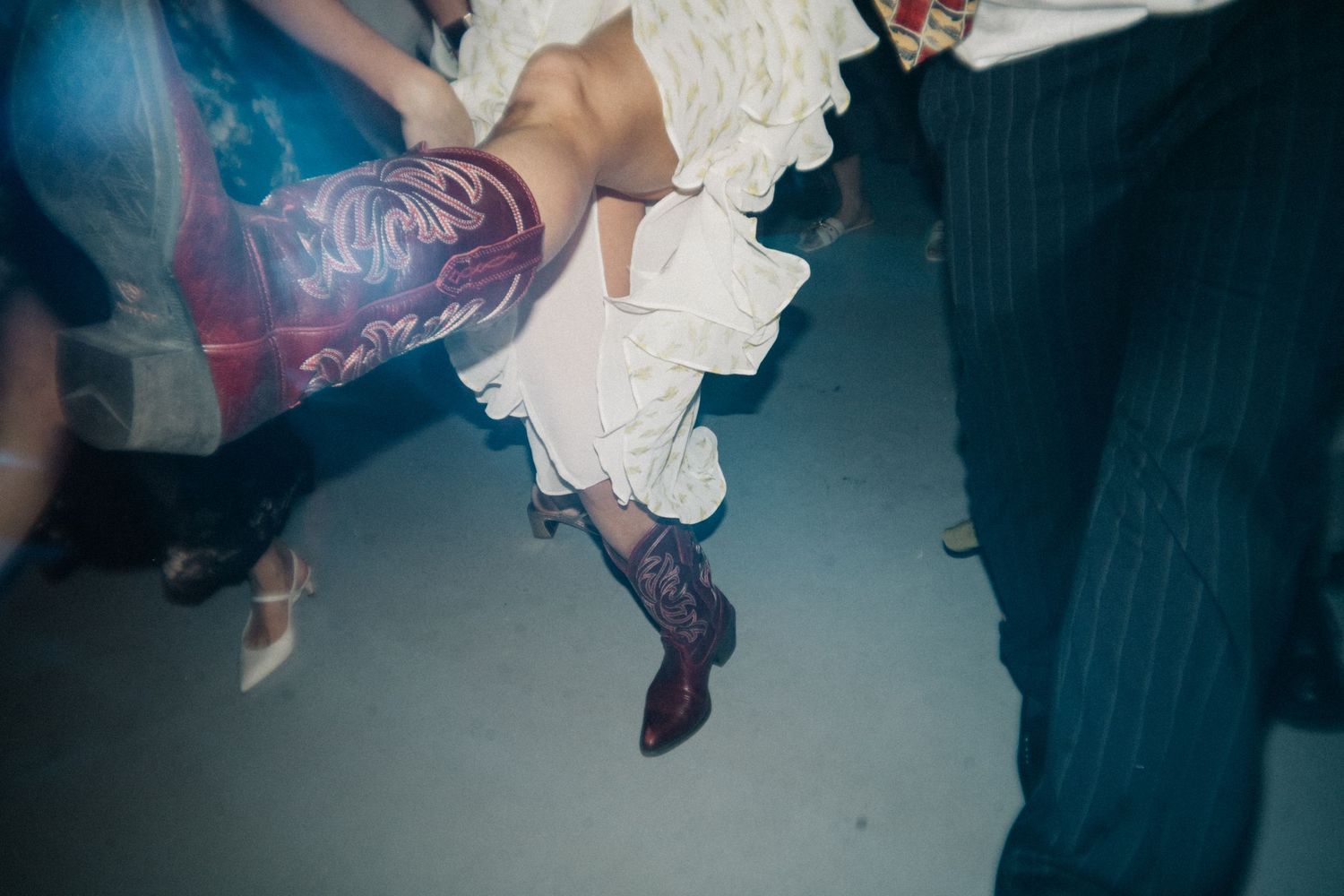 Guests dancing, seen at leg level: patterned boots, a white ruffled skirt and striped trousers.