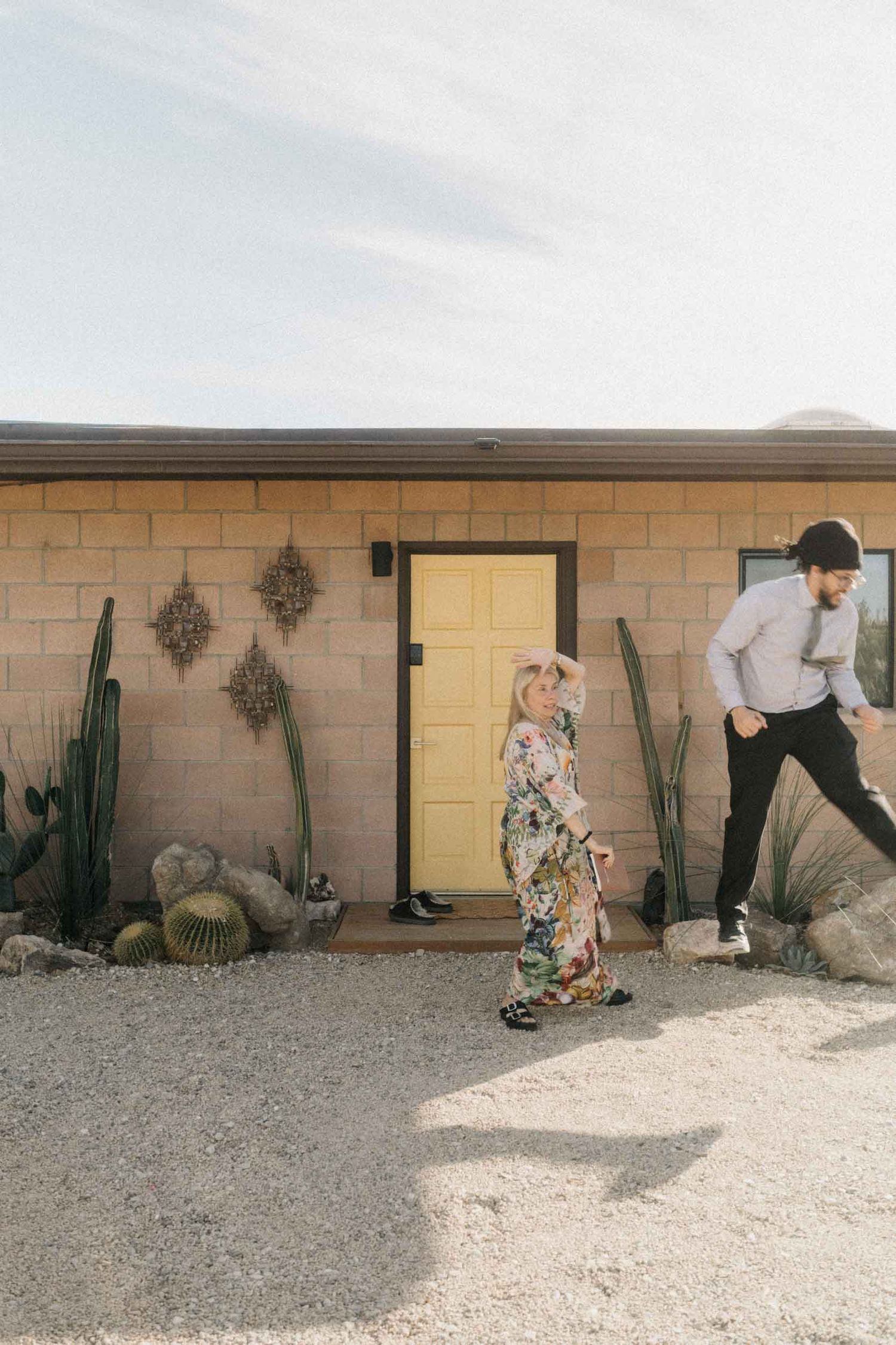 Two people walking near a small desert house with a yellow door and cacti