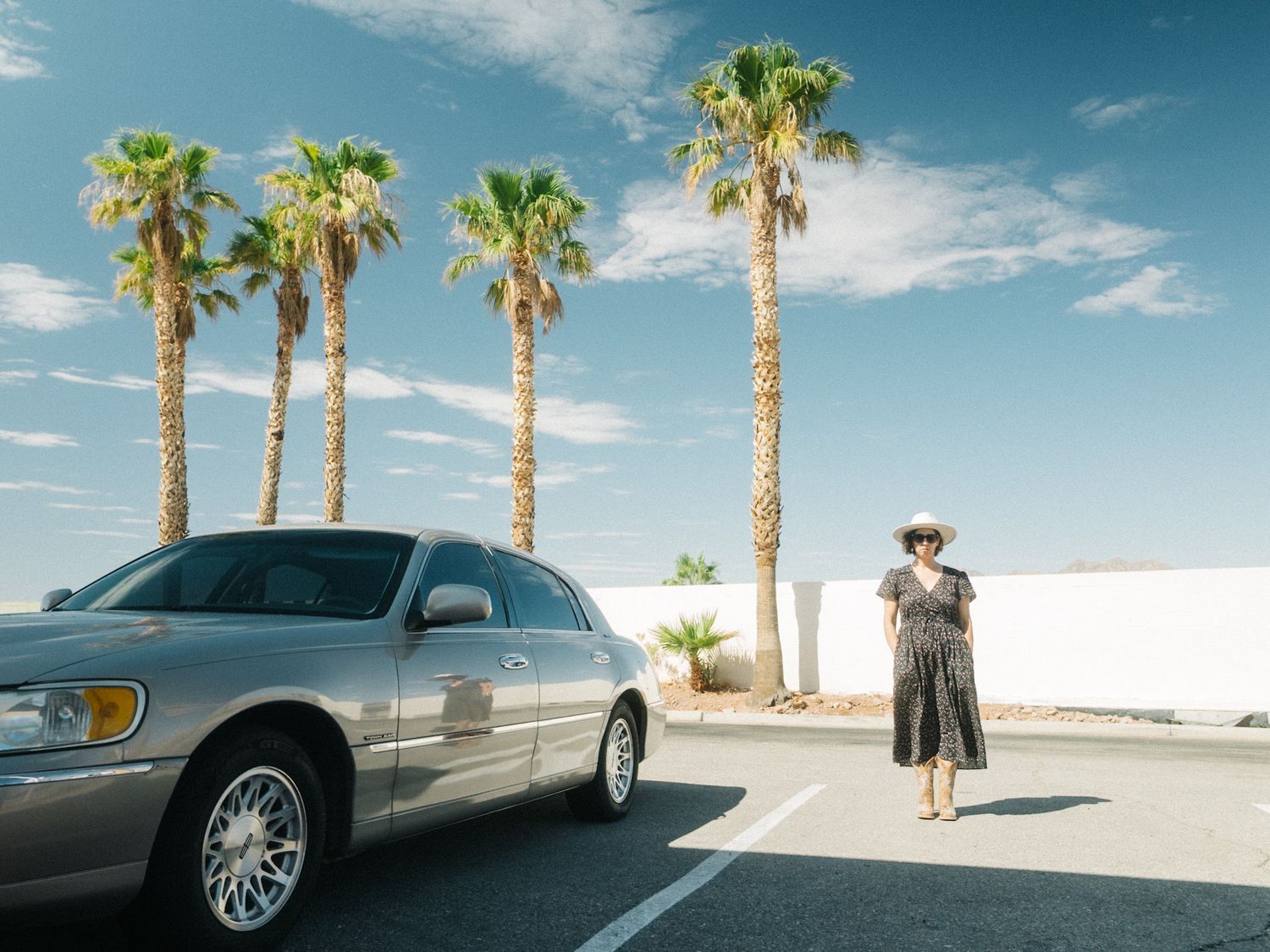 Documentary travel photography in Palm Springs, California — woman in a summer dress walking past a vintage car beneath tall palm trees.