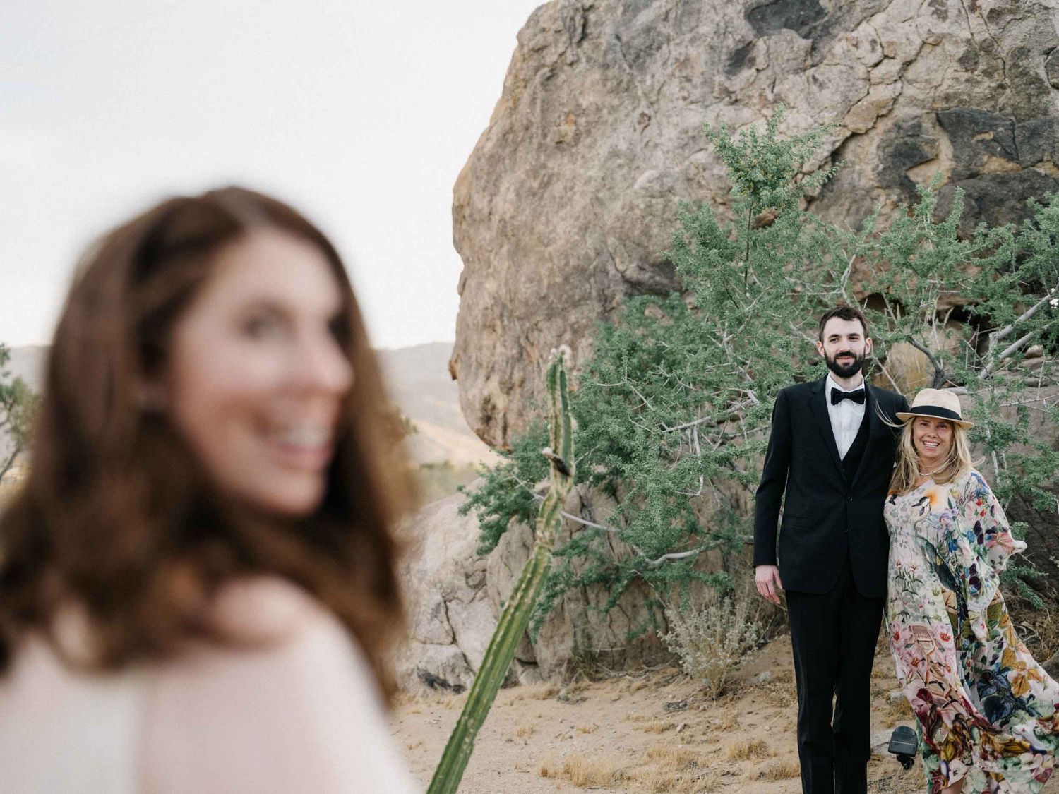 Man in a tuxedo and woman in a floral dress beside desert rocks, blurred portrait in foreground