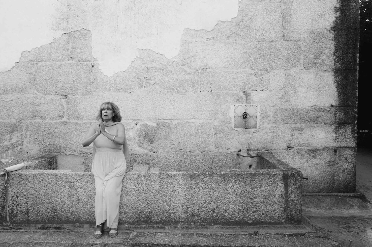 Black and white photo of a woman in light clothes standing with her hands together in front of a stone wall and trough.