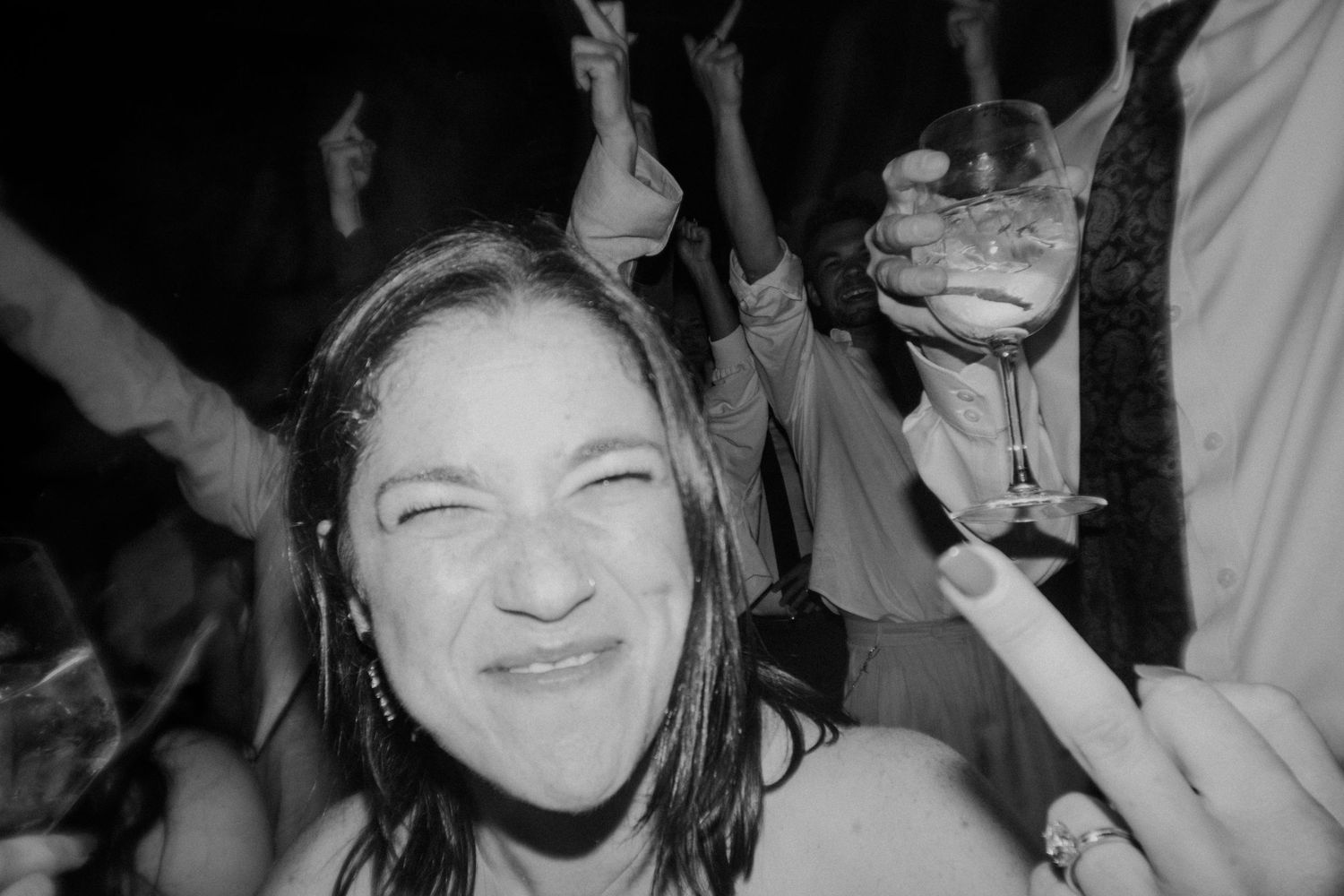 Black and white photo of a woman squinting with a big grin, with guests and a raised glass behind her.